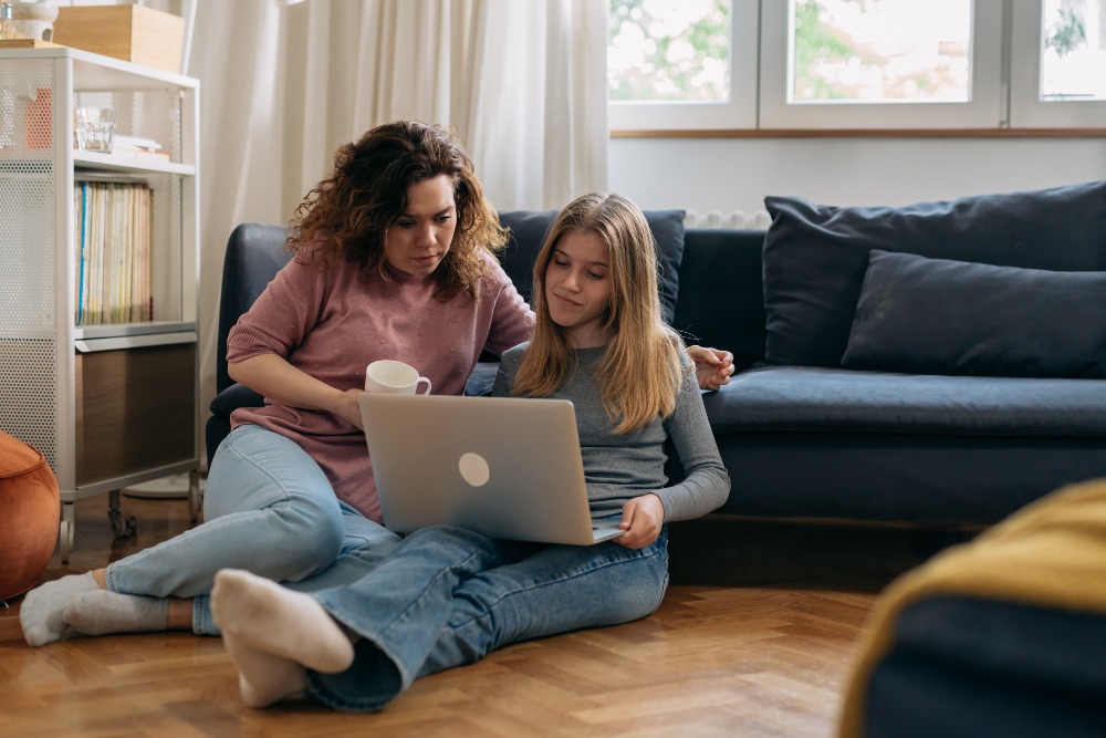 Two people sitting on the floor in a cosy living room, looking at a laptop together. One person holds a cup of tea while the other focuses on the screen, with both appearing engaged in what they are viewing. The scene is relaxed and homey, with soft lighting and a comfortable atmosphere.