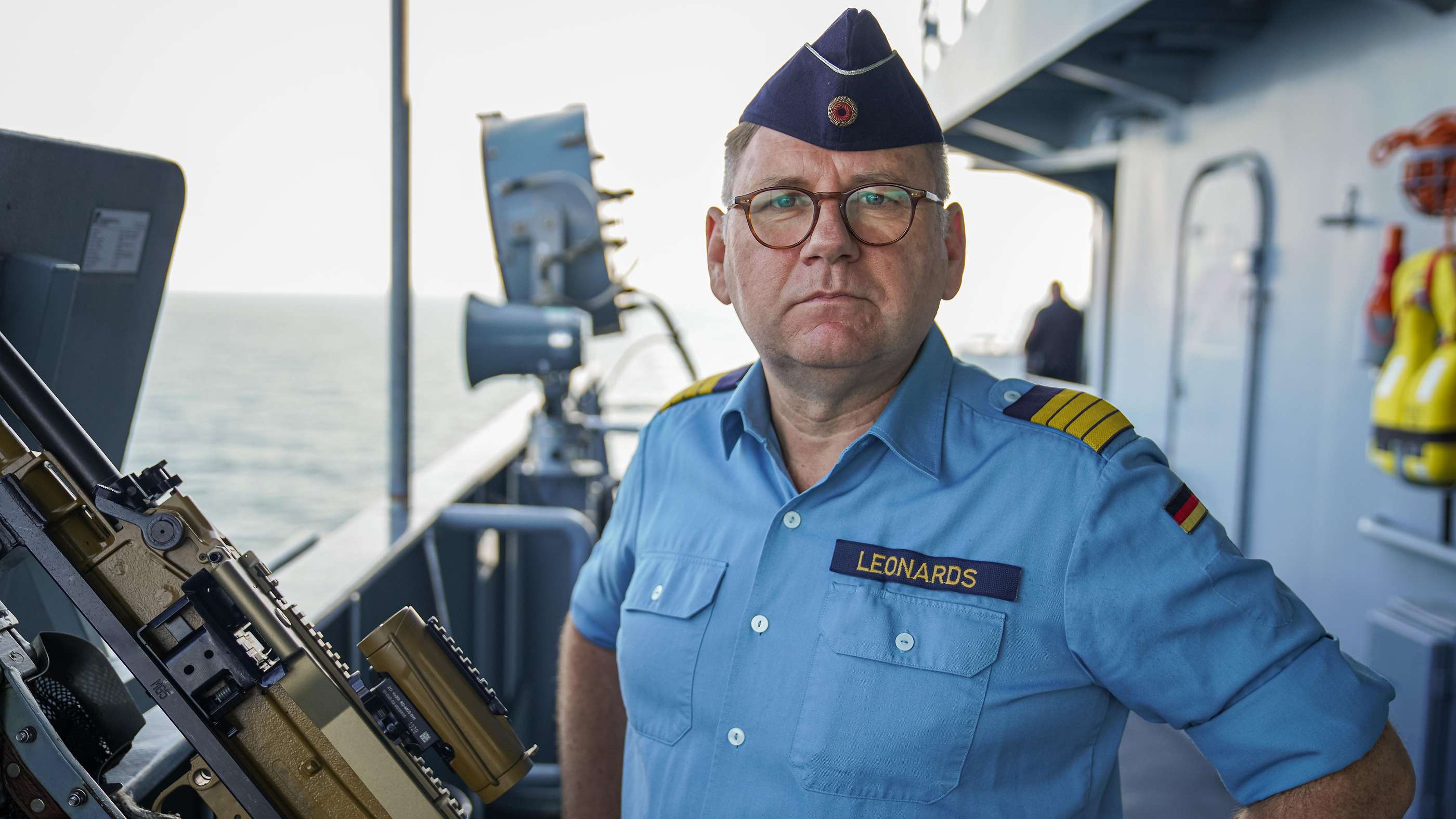 Ein Mann in Uniform steht auf einem Schiffsdeck. Neben hier hängt am Rande des Decks eine Waffe. 