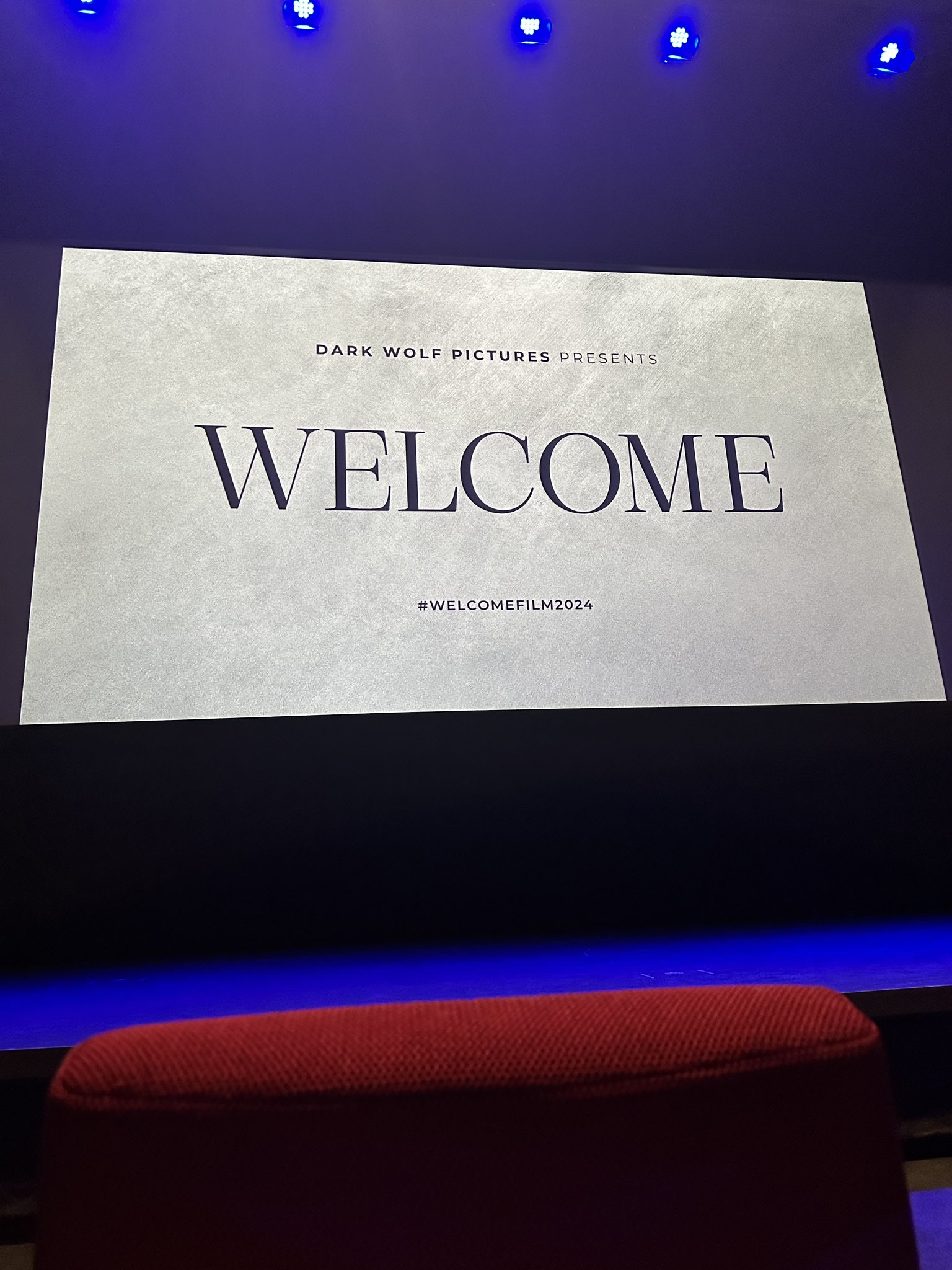 A movie theatre screen with black text on a white background. The background is slightly textured, almost like a paper with some wrinkles on it. The text reads “dark wolf pictures presents” at the top, “Welcome” in huge letters in the centre of the screen, and “#welcomefilm2024” in smaller font on the bottom.