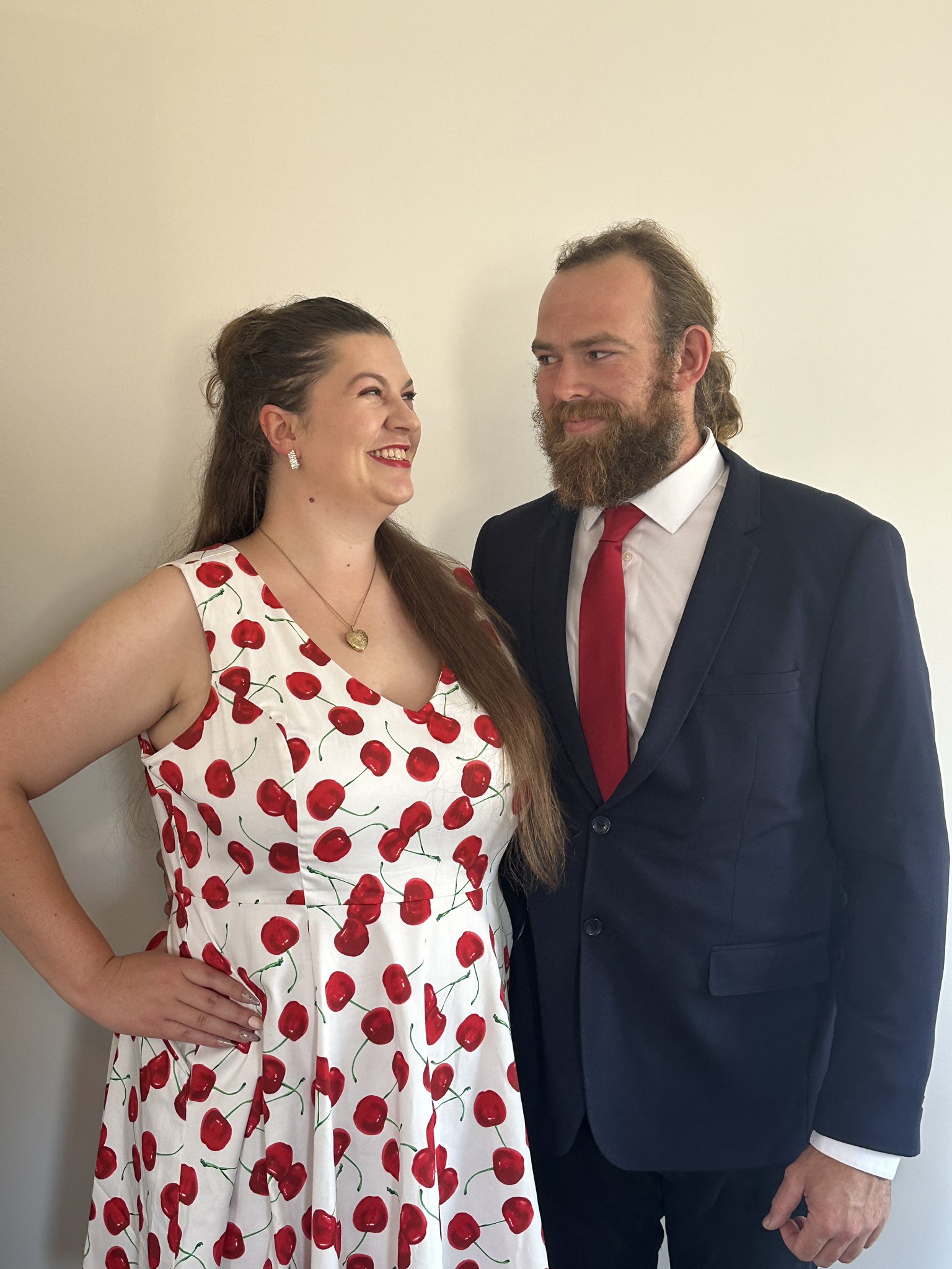 Lauren and her partner Hudson are dressed fancily and gazing lovingly into each others’ eyes. Lauren wears a white dress with cherries all over it, sparkly earrings, and a gold heart shaped locket. She has very long dirty blonde hair that is half up and half down. Hudson wears a blue suit with white shirt and red tie. He has dirty blonde hair in a ponytail with a big scruffy beard and moustache. The white and red of their outfits match. Hudson is a little taller than Lauren in this photo because Lauren is wearing high heels, but they are not in frame.