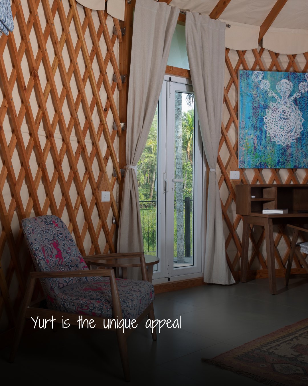 Cozy interior of a yurt featuring a patterned armchair, wooden lattice walls, and a glass door with beige curtains overlooking greenery.