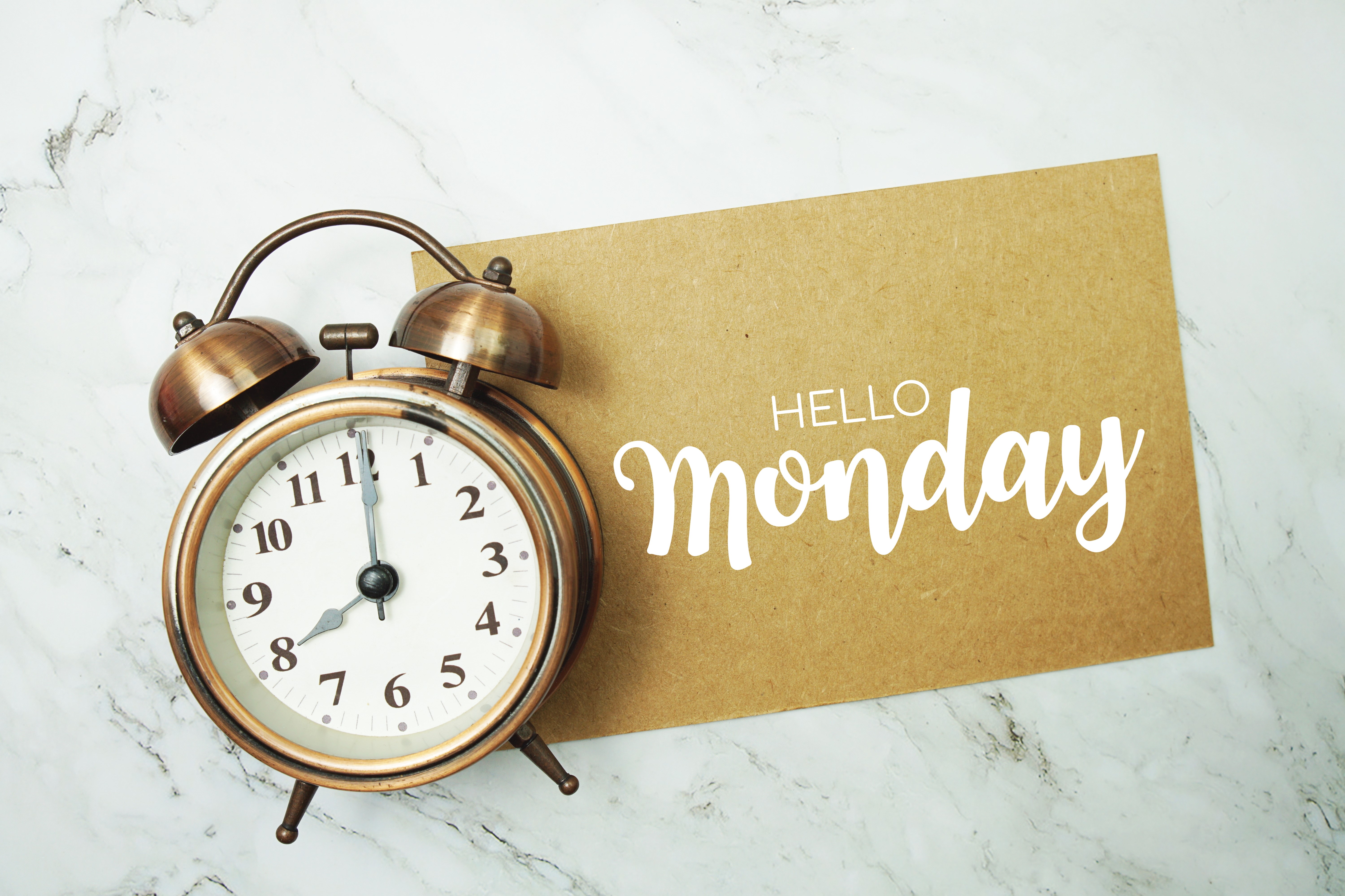 Vintage brass alarm clock showing 7:00 on a marble surface next to a brown card with 'Hello Monday' in white script.