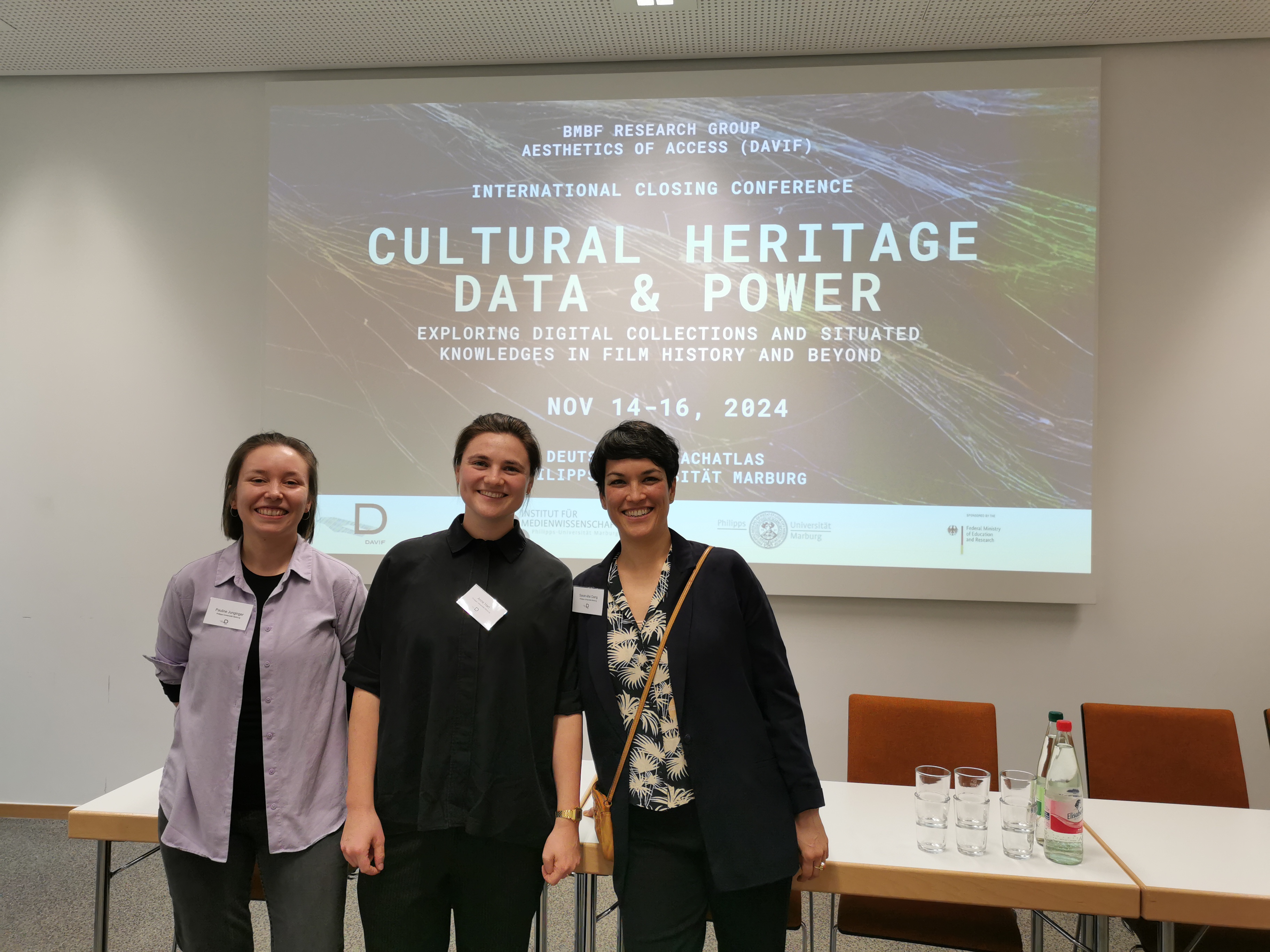 Pauline Junginger, Anne Hart & Sarah-Mai Dang standing in a conference room in front of slides with the poster of the DAVIF closing conference. 