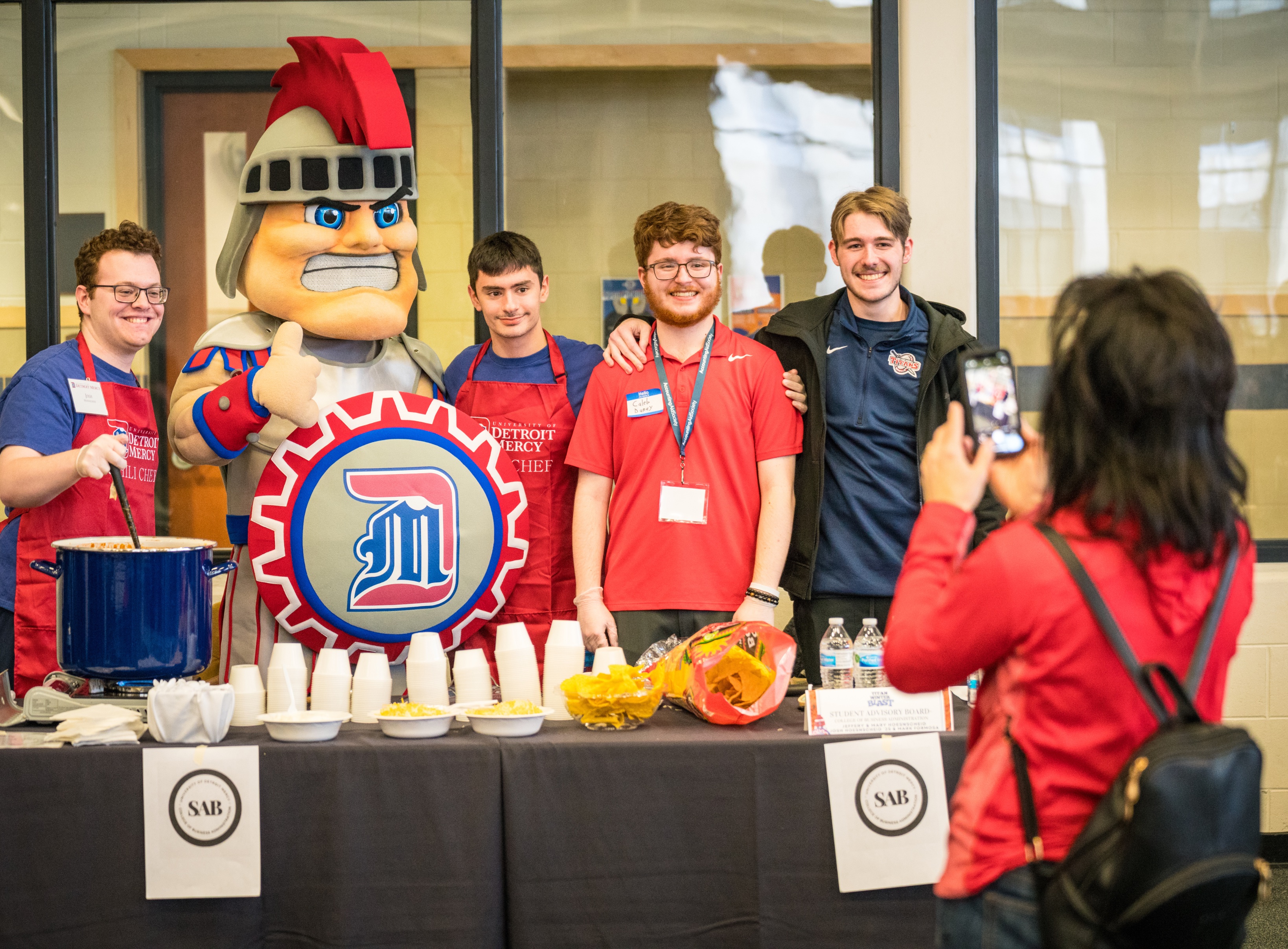 Four students and Tommy Titan pose as they have their photo taken at a chili cook-off table inside of the Student Fitness Center.