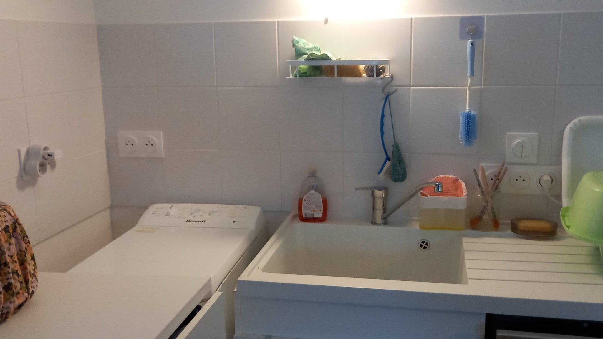 Photo. À gauche une machine à laver top et 3 prises électriques a environ 20 cm au dessus de celle-ci. A droite l'évier avec 3 prises et des interrupteurs à environ 30 cm du robinet