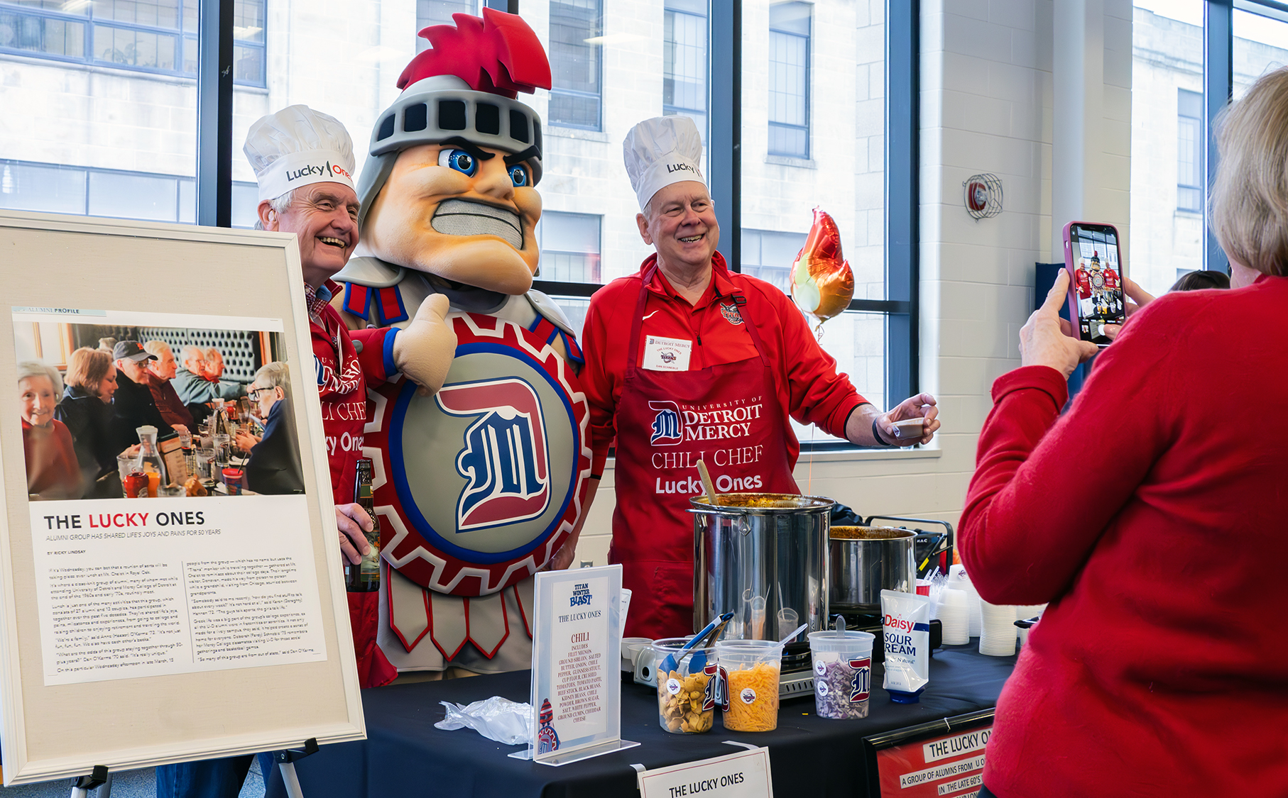 A photo of Tommy Titan posing with two Titan alumni during the Titan Winter Blast inside of the Student Fitness Center. A posterboard next to them reads, THE LUCKY ONES.