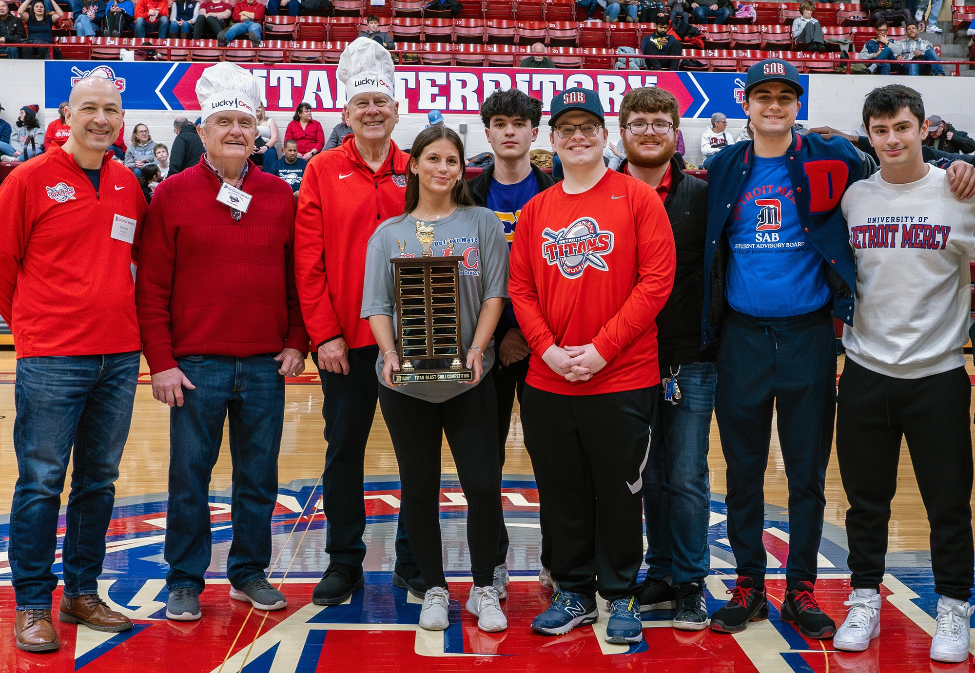 Nine people, some wearing hats and one holding a trophy, pose for a photo inside of Calihan Hall in front of a TITAN TERRITORY sign.