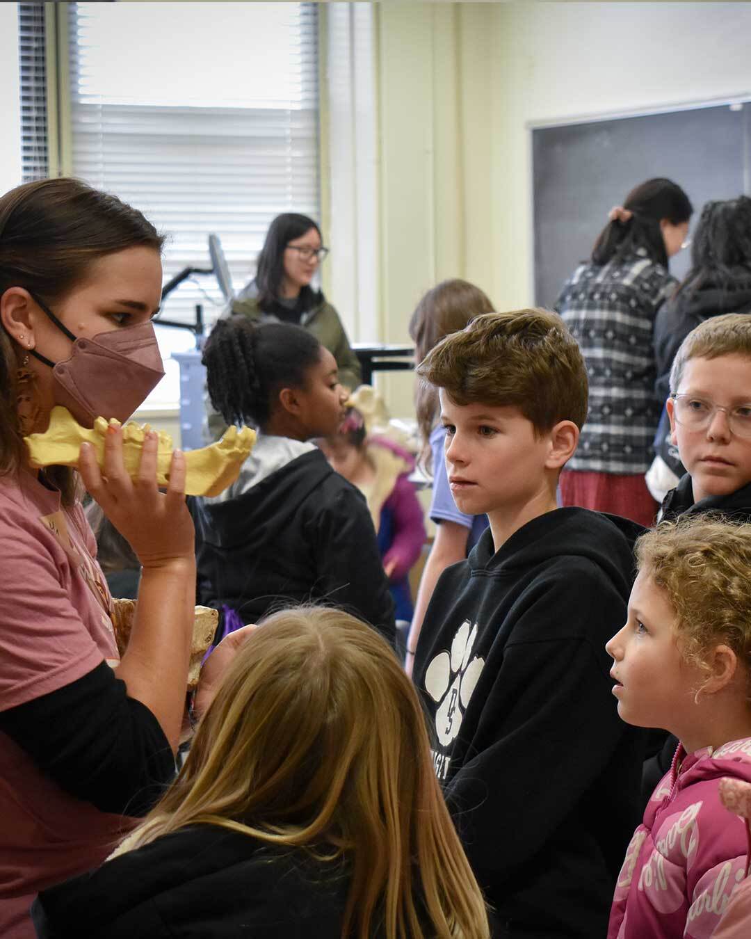 A woman wearing a mask and a pink t-shirt explains about a small fossil jaw she’s holding to two young boys and two young girls.