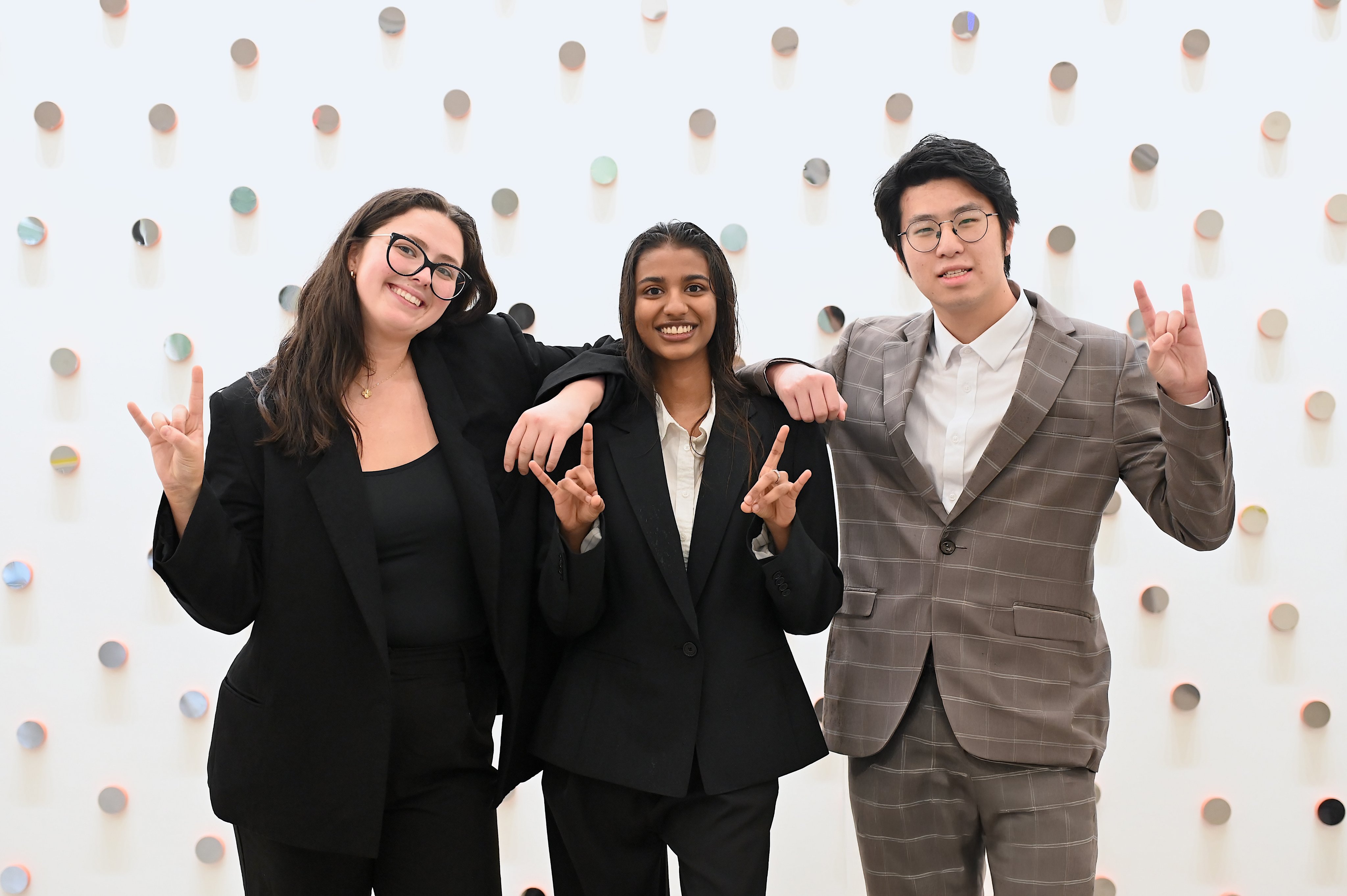 Three students stand in front of a wall studded with small circular mirrors. All three are wearing suit jackets and smiling while holding up the Hook ’em Horns sign with their hands. The left is a woman with brown hair wearing black-framed glasses. In the middle is a woman with dark brown hair. On the right is a man with black hair wearing wire-rimmed glasses.
