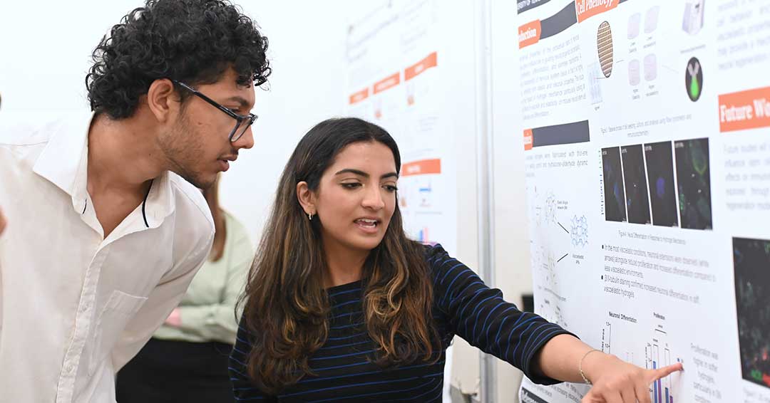 A young woman points to data on her research poster as she explains the work to an onlooker.