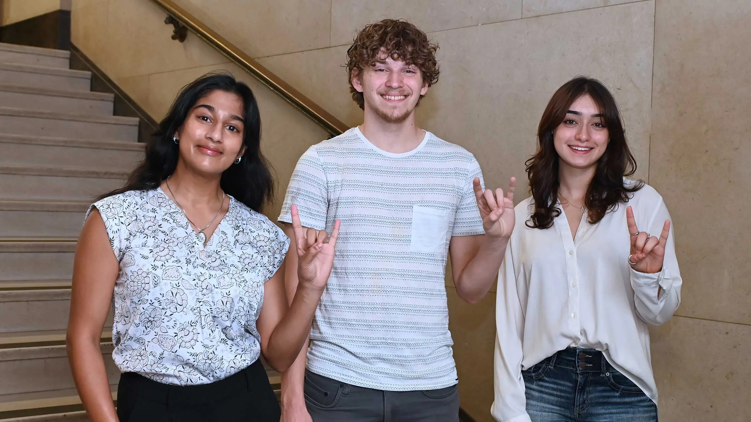 Three smiling students give the Hook'em Horns sign in a stairwell on campus.