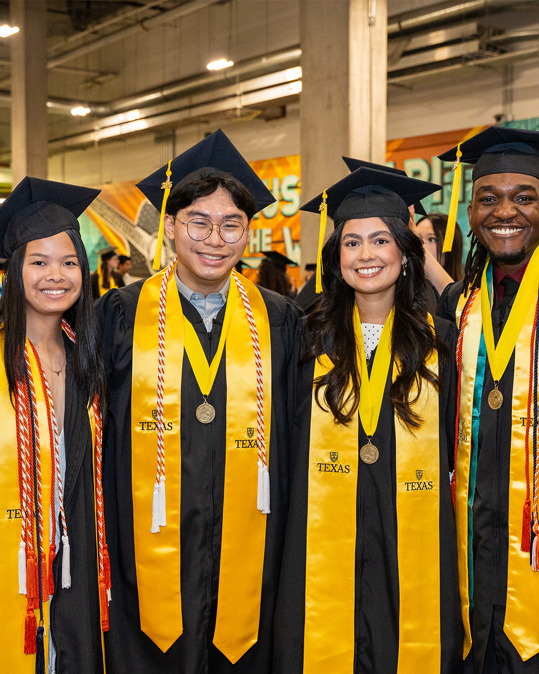 Four graduates stand shoulder to shoulder smiling. On the left is an Asian-American woman, to her left is an Asian-American man, to his left is a LatinX woman’s and to her left is a Black man wearing a First-Gen Grad stole.