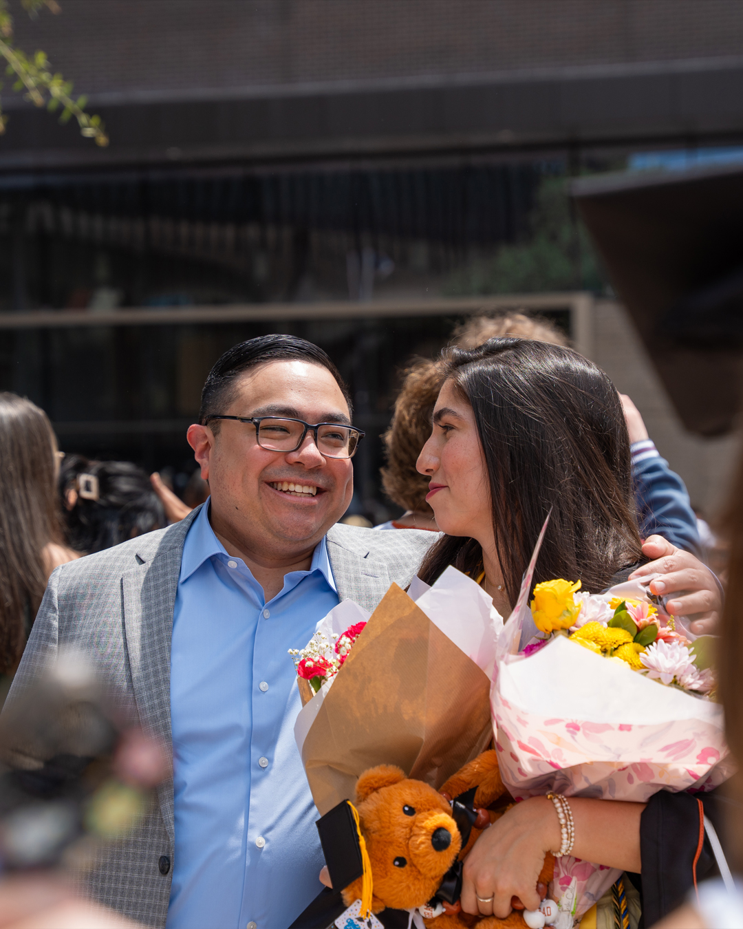 A middle-aged LatinX man smiles and looks at the woman he is hugging with his left arm. The young woman smiles and looks back at him. She is carrying two bouquets of flowers and a burnt orange teddy bear that is wearing a graduation cap.