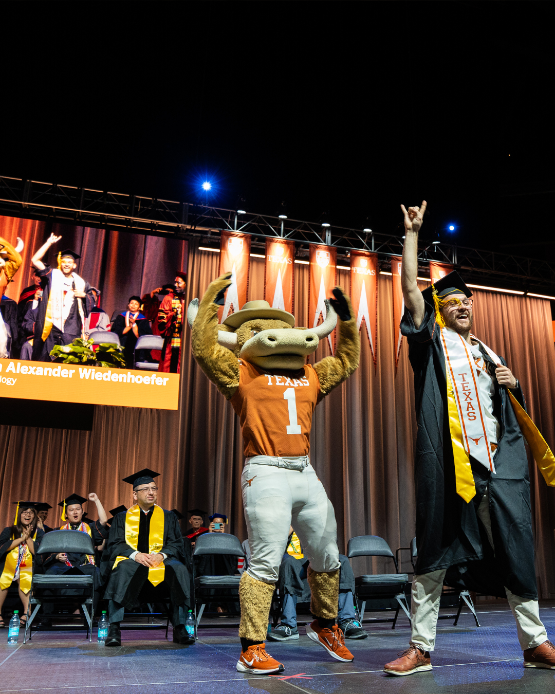 A bearded man wearing a Texas Mascot stole cheers as he holds his right hand above his hand with the Hook’Em Horns sign. Behind him, to his right is the mascot Hook’Em with both hands raised. Behind both of them to their right are several people seated in graduation garb cheering and smiling. A video screen is just visible above the seated people and reads “Alexander Wiedenhoefer” and shows the first man from a different angle.