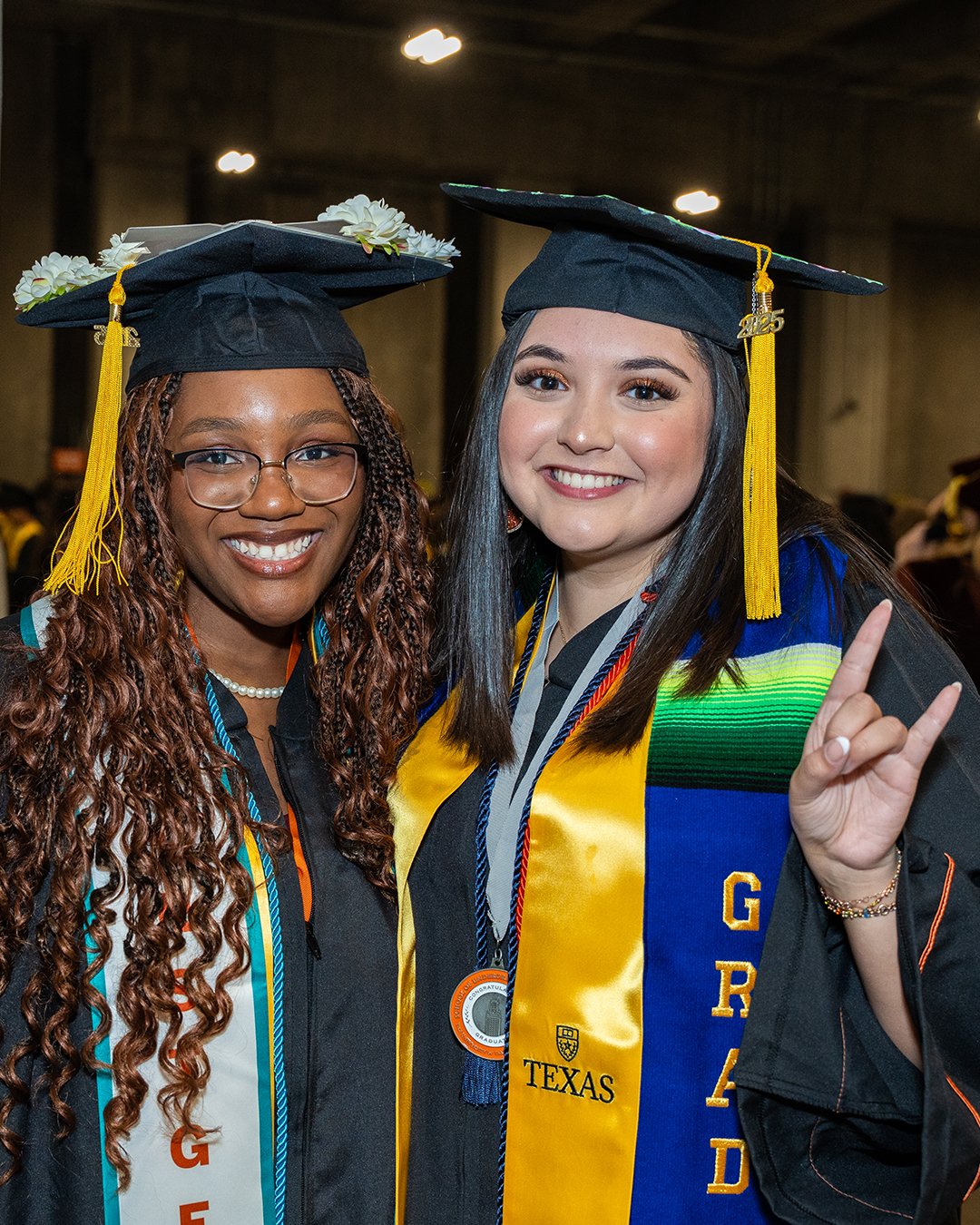 Two women wearing graduation garb smile and huddle together. The woman on the left is Black and wearing a First-Gen Student stole. The woman on the right is LatinX and is wearing a LatinX Grad stole and holding up her left hand in the Hook’Em Horns sign.
