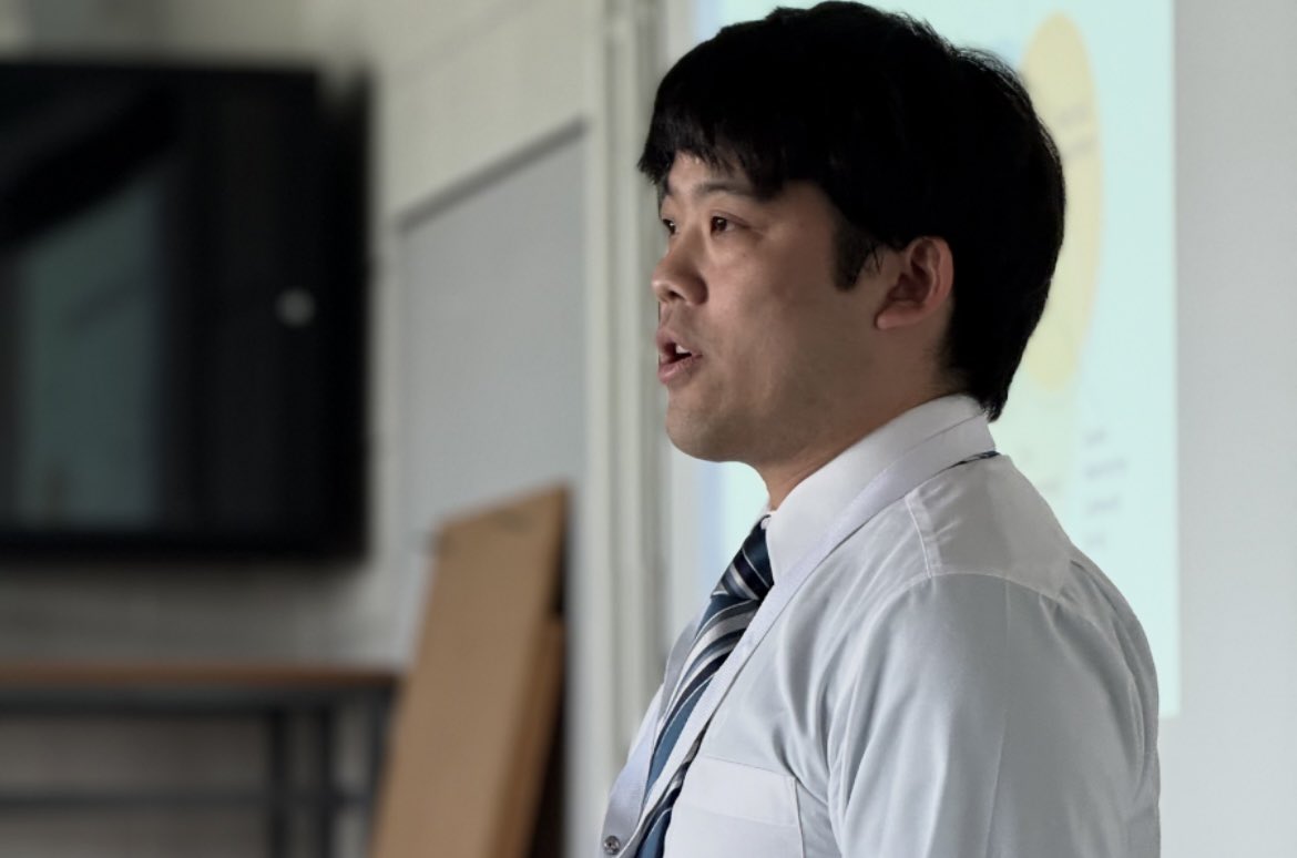 A man in a white shirt and striped tie is speaking in front of a classroom, facing to the right. He appears to be giving a presentation, with a blurred projection in the background and a serious expression on his face.