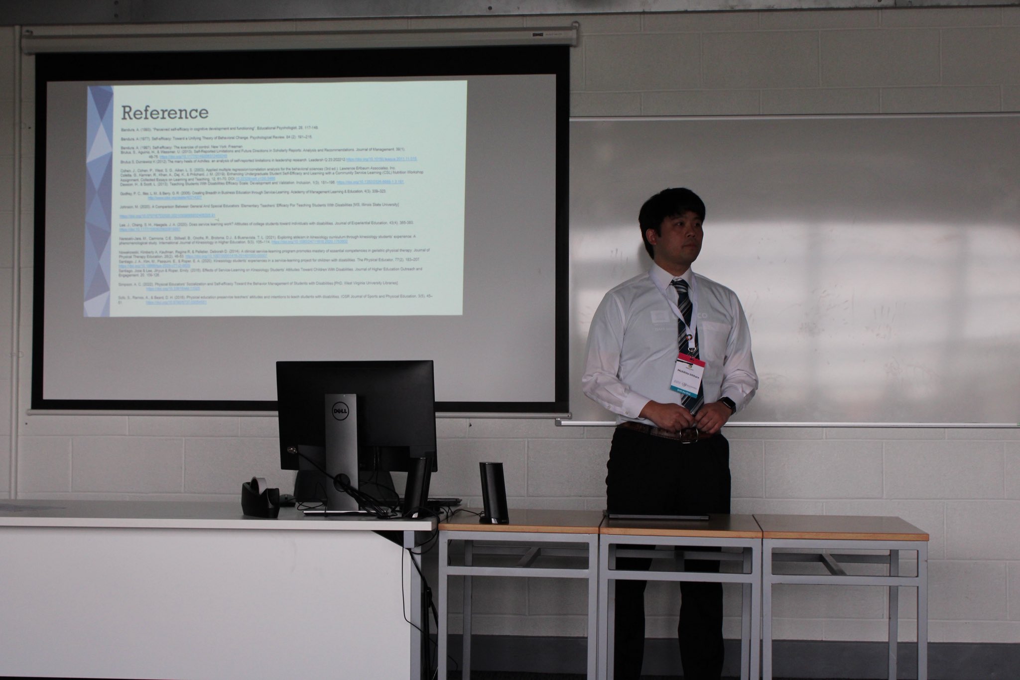 The same man is standing in front of a projector screen displaying a slide titled “Reference” during a presentation. He is wearing a conference name badge and is positioned next to a computer on a desk. The classroom has white walls and a large whiteboard.