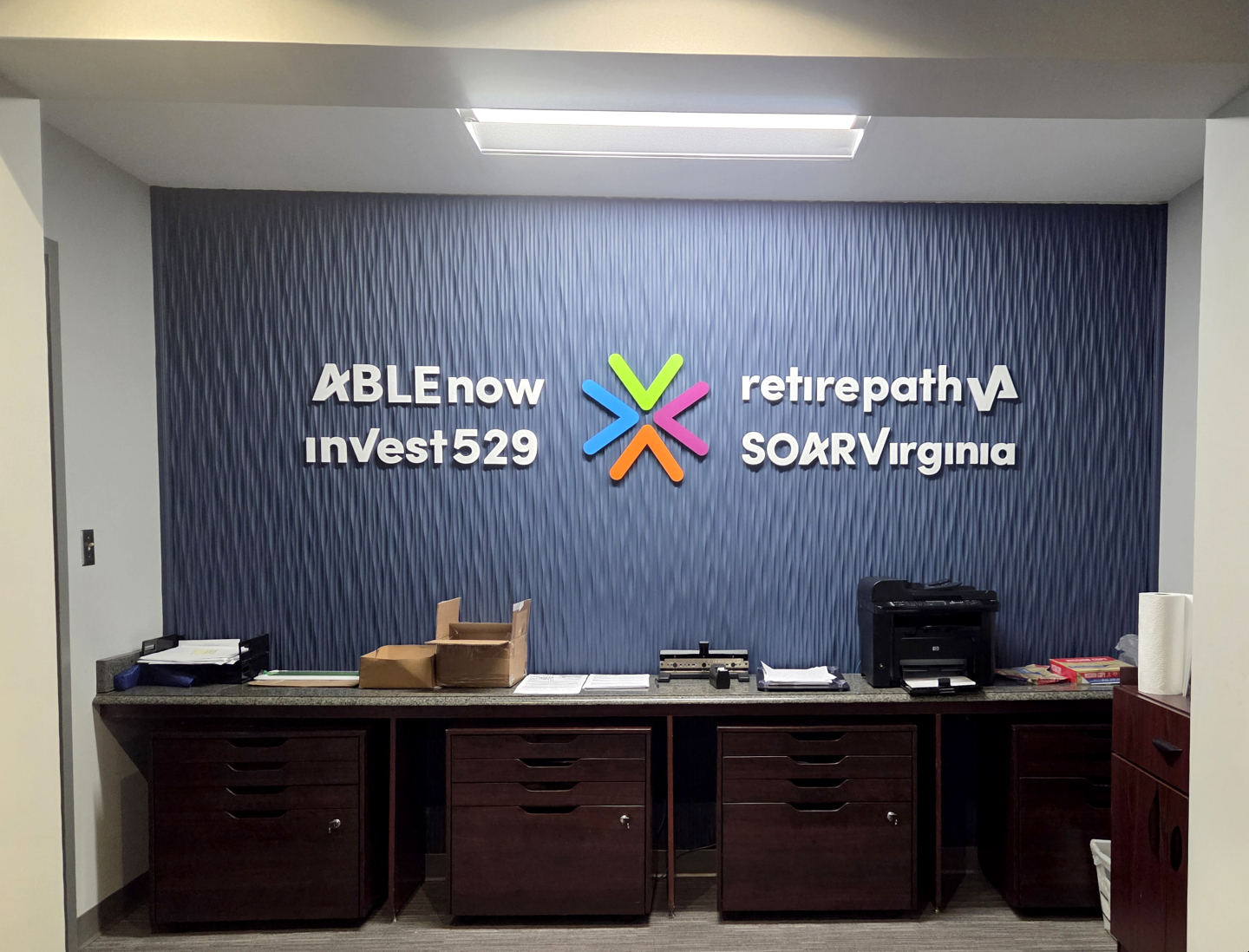 Office wall with logos for ABLEnow, inVEST529, retirepathVA, and SOAR Virginia, above a counter with office supplies and a printer.