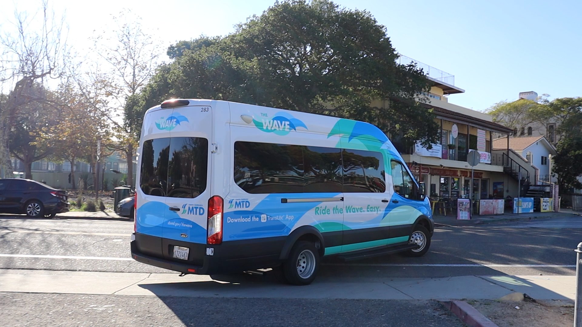 A blue-and-teal Santa Barbara MTD “Wave” microtransit van drives through a sunny neighborhood intersection, with trees and storefronts in the background. The vehicle branding reads “Ride the Wave. Easy.” as the van turns onto the street in bright afternoon light.