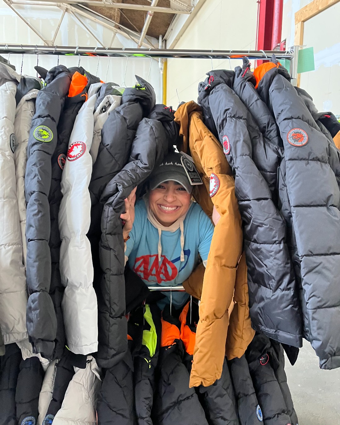A person playfully peeks through a rack of hanging puffy coats, including several Canada Goose jackets, in a warehouse or store setting.