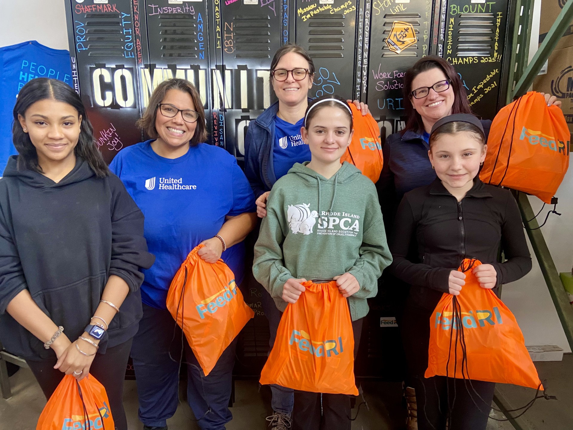 A group of six people smiling and holding orange FeedRI bags. Behind them is a wall with various writings on a chalkboard, including "COMMUNITY" in large letters. One person is wearing a blue United Healthcare shirt.