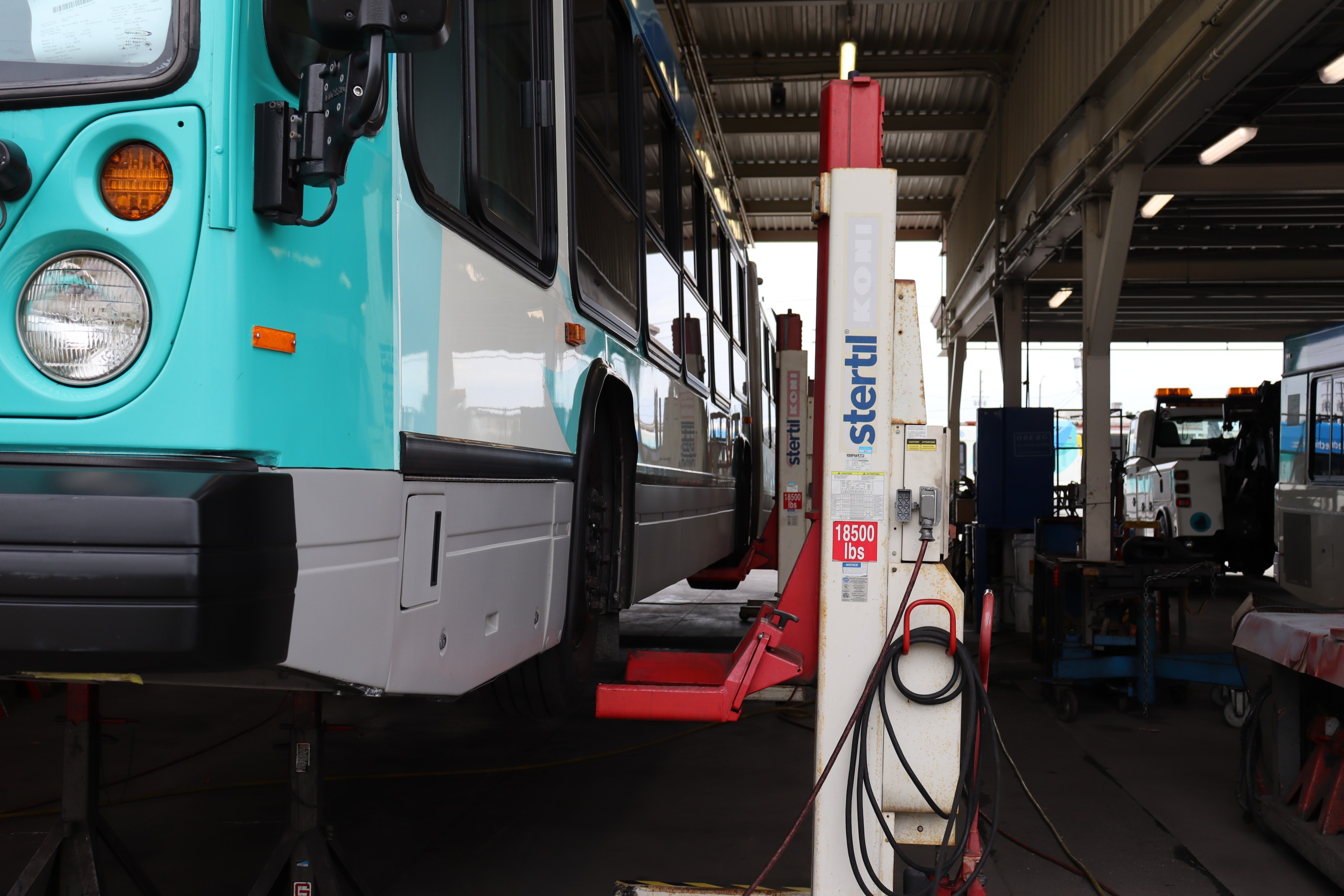 An MTD bus lifted high on a hydraulic lift inside a maintenance bay while technicians work underneath it. The large vehicle is elevated above the shop floor with tools and equipment visible, capturing a behind-the-scenes moment of routine maintenance and inspection.