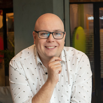 A smiling bald man with glasses rests his chin on his hand. He wears a patterned white shirt with a pocket. The background includes a window and decor.