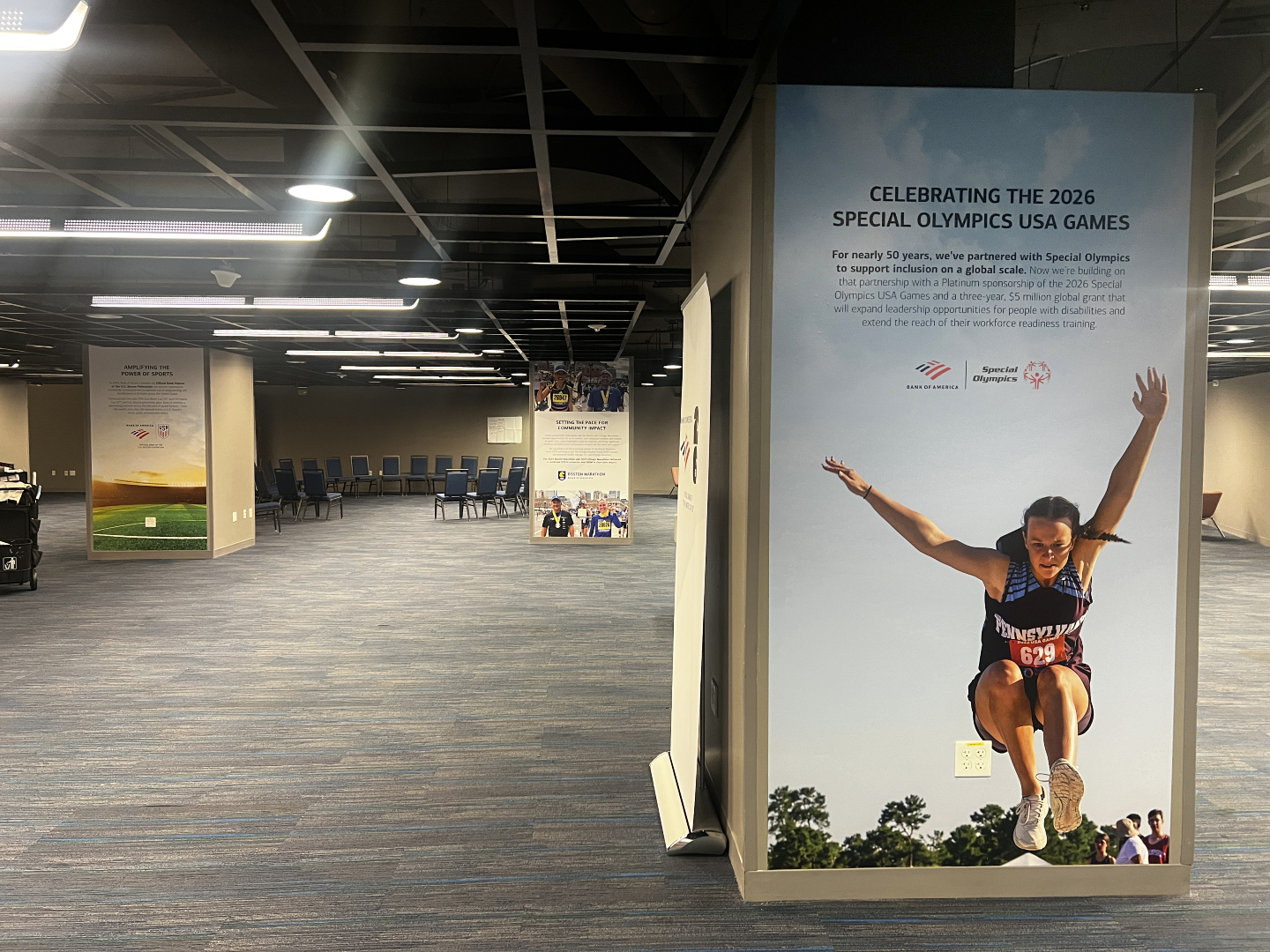 Event hall with large Special Olympics USA Games 2026 wall poster of a female athlete mid-jump and empty stacked chairs.