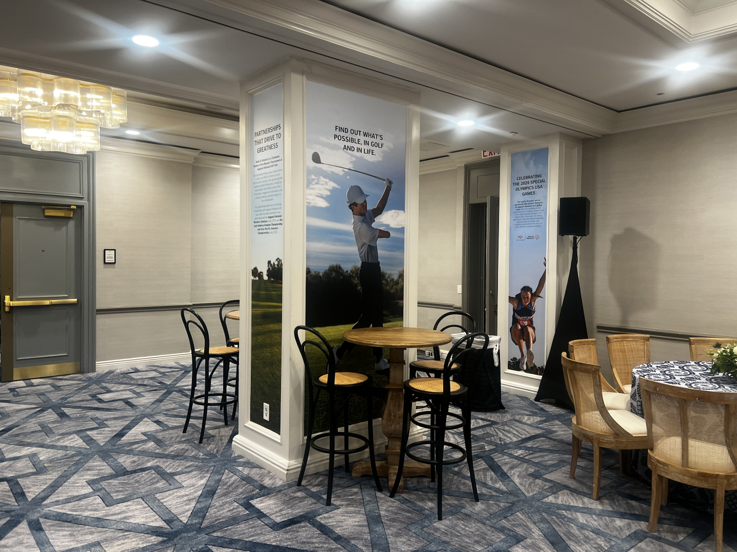 Hotel meeting lounge with tall round table and stools beside pillars displaying large golf and athlete posters.