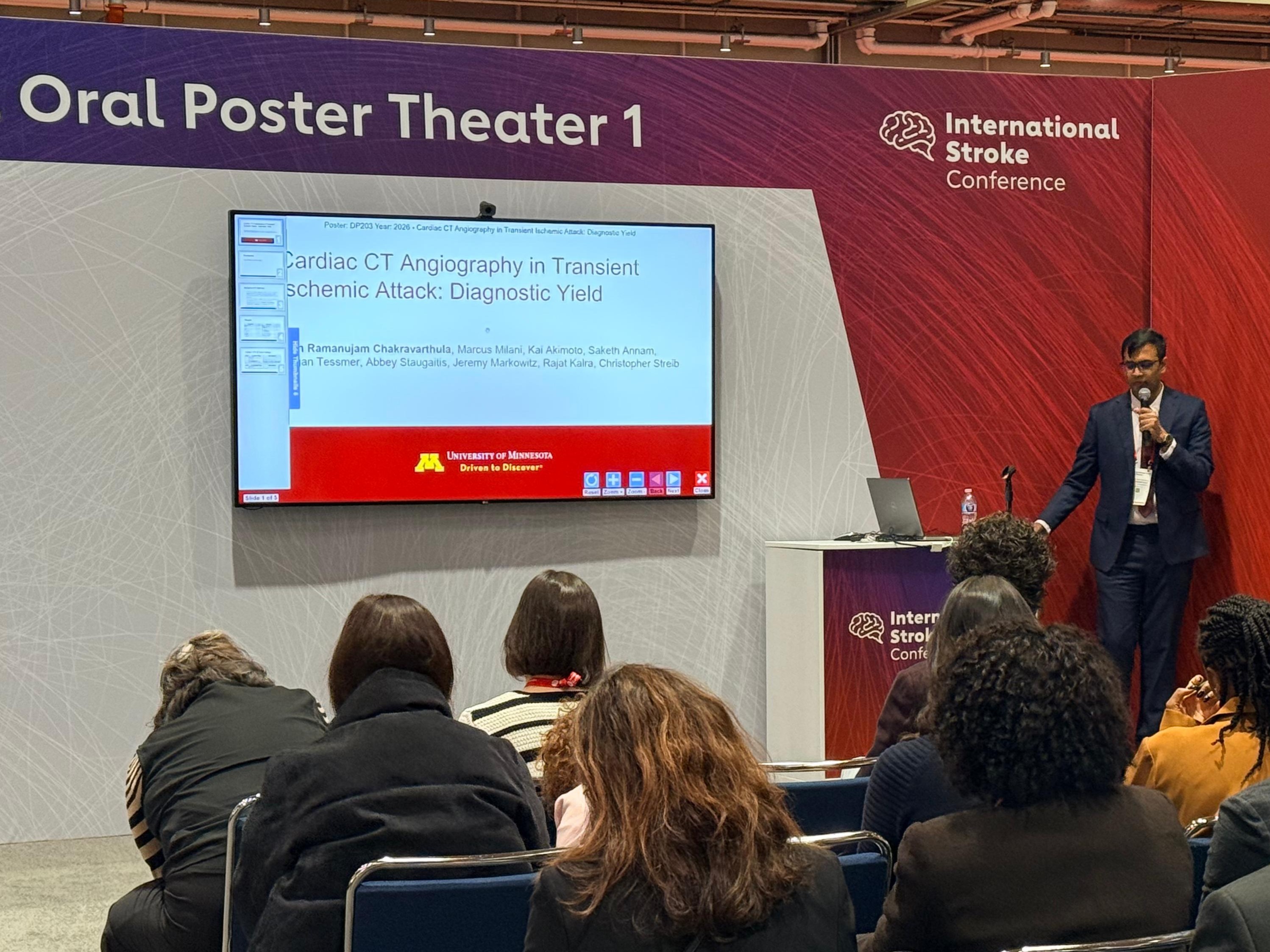 Dr. Chakravarthula stands at a podium with a microphone to present his slide deck. He is in front of a background marked with the International Stroke Conference logo and "Oral Poster Theater 1." About 10 people are pictured in the audience.