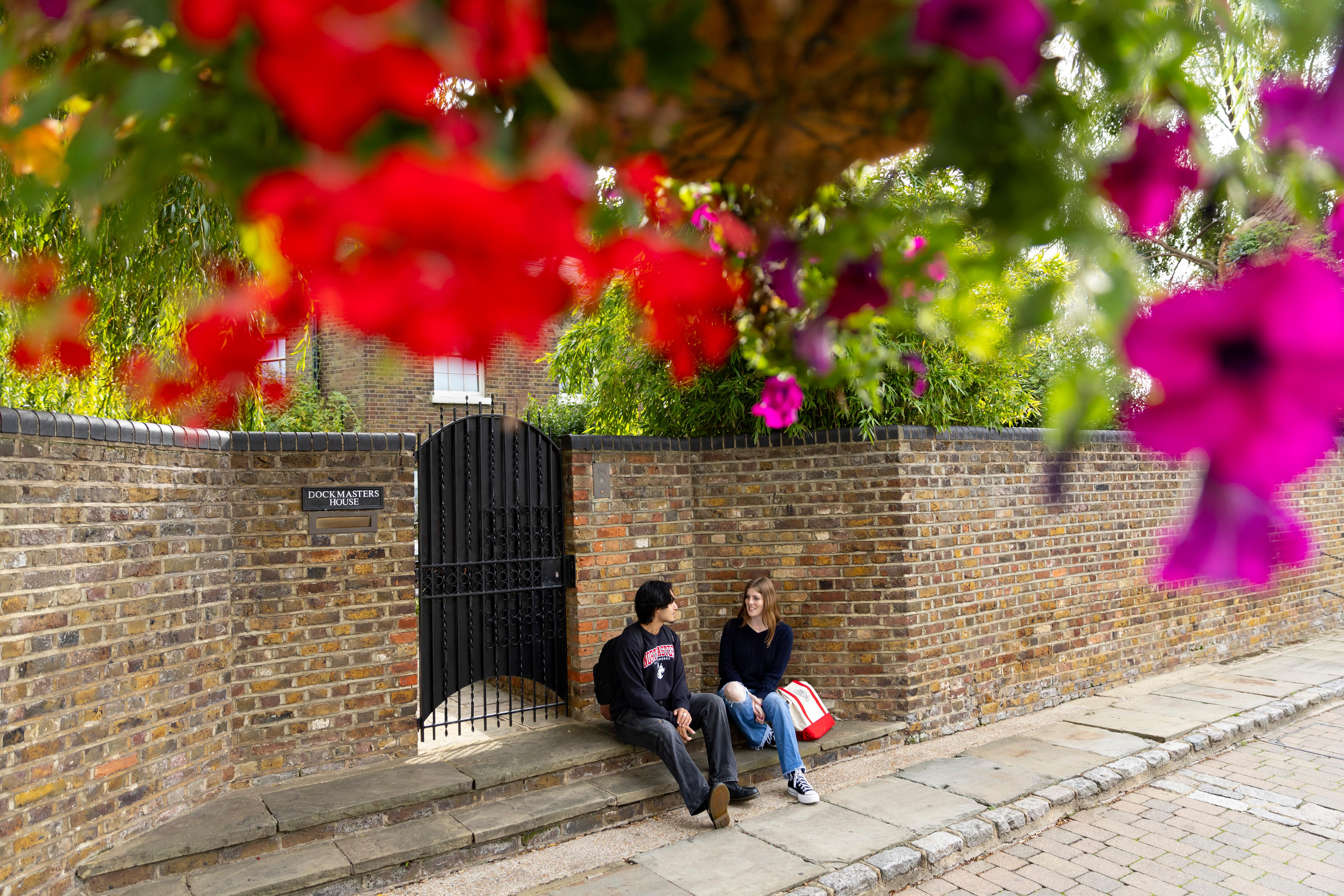 Two students sit on stone steps beside a brick wall under hanging flower baskets. A gate and a sign reading \"DOCKMASTERS HOUSE\" are visible on the wall. The two individuals appear to be conversing.