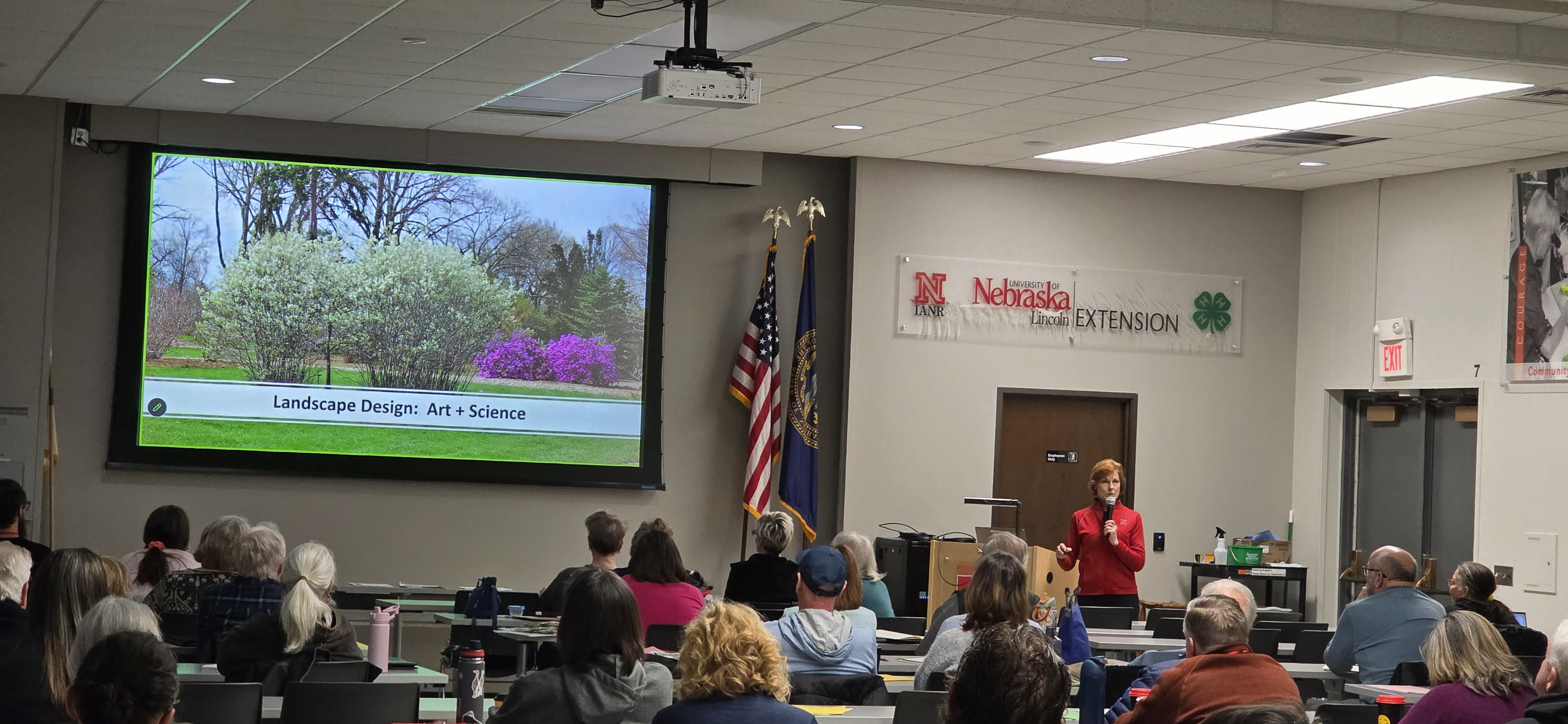 Kim Todd, in a red shirt speaks to a seated audience in a University of Nebraska–Lincoln Extension meeting room. A large screen beside her displays a slide reading, “Landscape Design: Art + Science,” over a photo of flowering shrubs and trees.