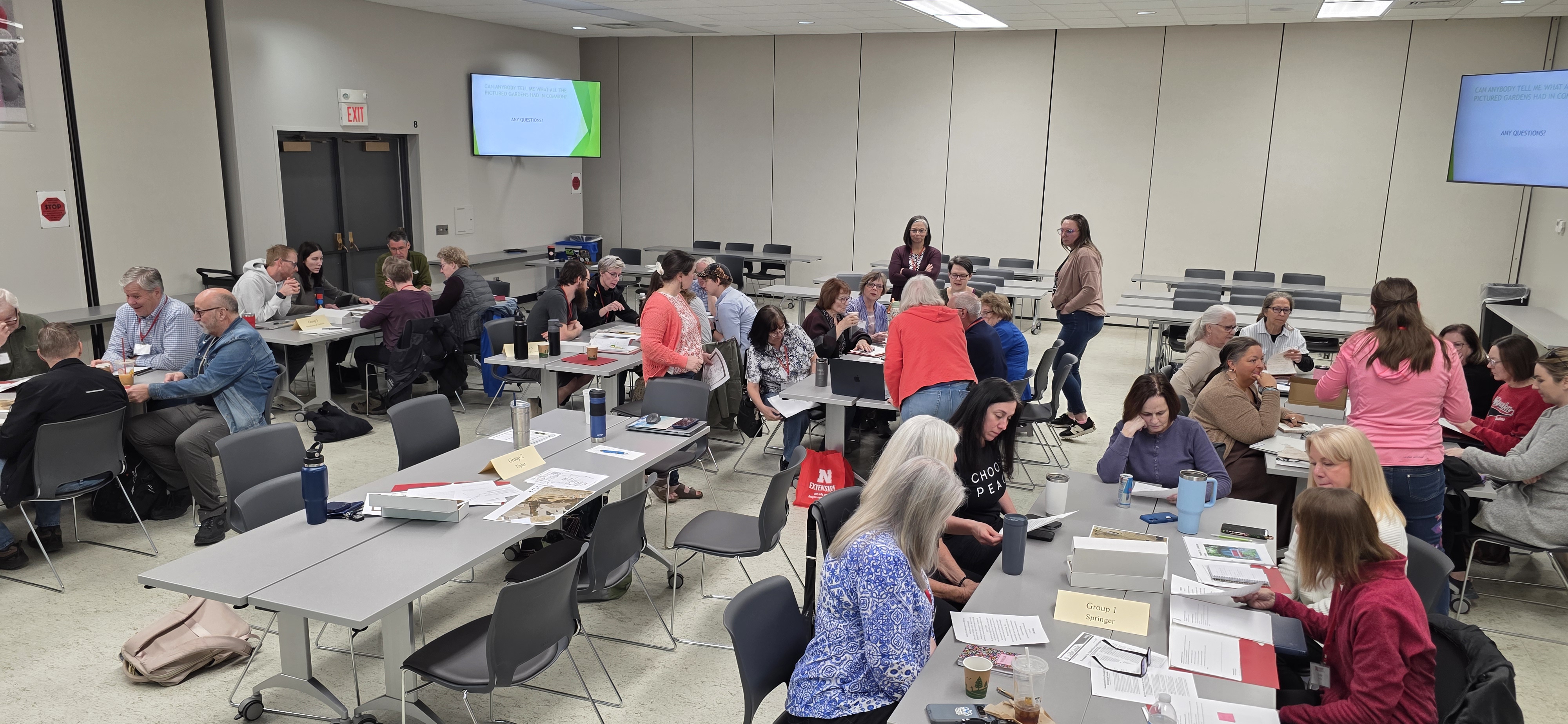 Extension Master Gardener Volunteer interns work in small groups around tables during a classroom activity, applying landscape design concepts from the previous week’s class. Papers, plans, and discussion materials are spread across the room as participants collaborate.