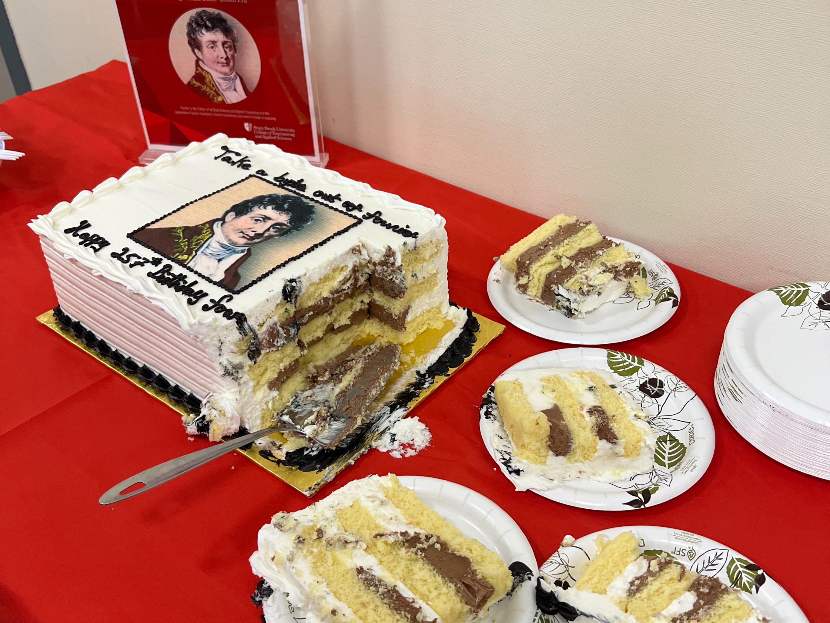 A partially eaten sheet cake with an edible image of Jean-Baptist Joseph Fourier on top. The cake is on a red tablecloth next to several paper plates with cake slices. A display in the background features a similar image of Fourier.