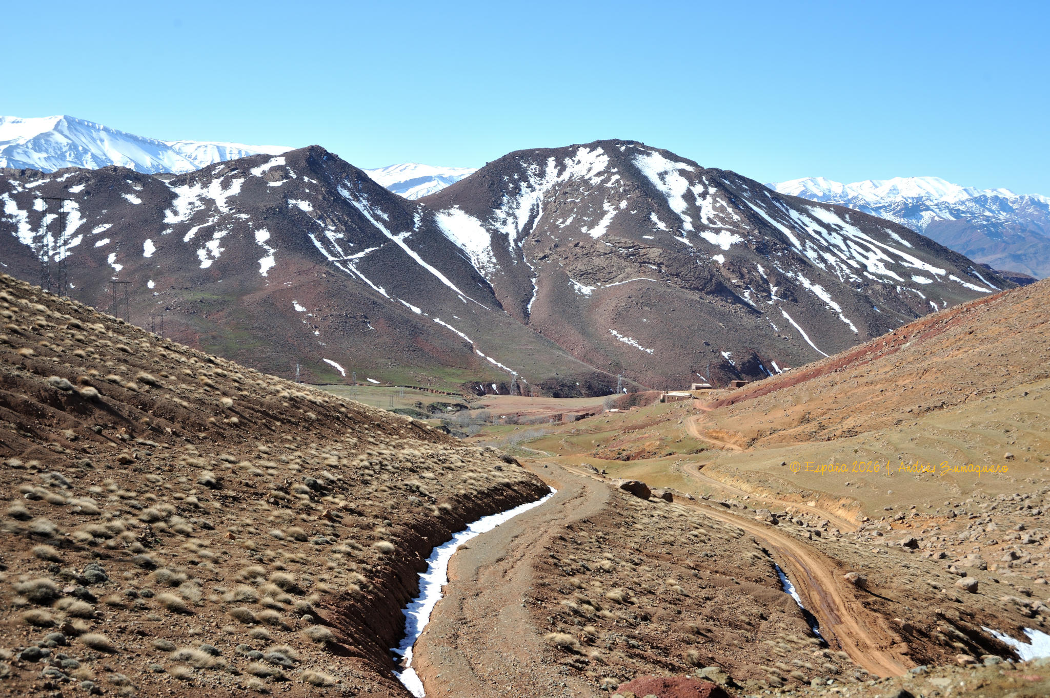 La parte superior de la imagen es de unas montañas, las más lejanas con nieve en las cumbres y las cercanas en la ladera, aunque no mucha. La mitad inferior de la imagen es de una ladera a cada lado de cuadro, un pequeño valle que, en su mediación, parece que tiene un cauce, tal vez de un torrente.