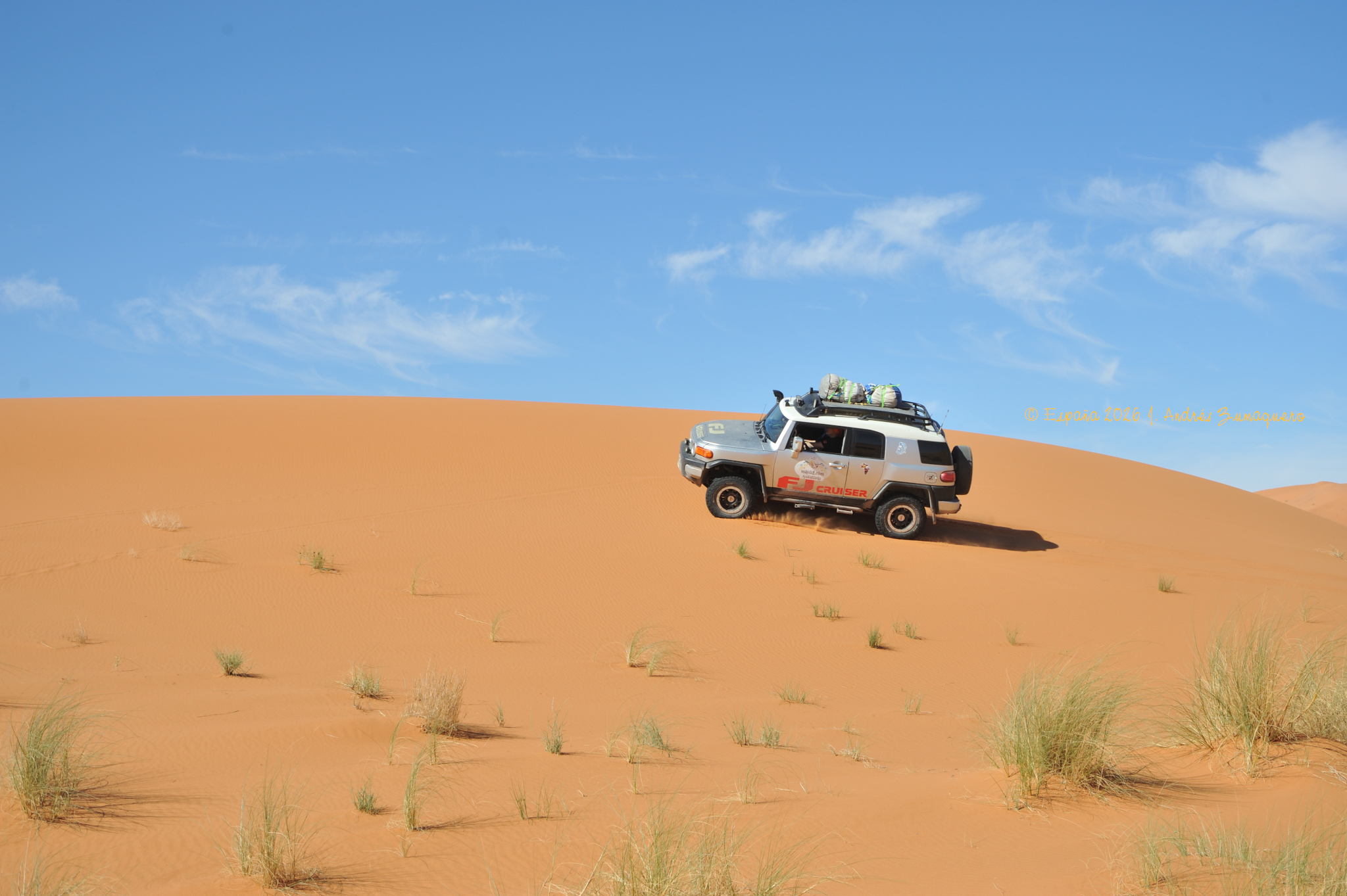 La imagen está dividida ente la arena de la duna, sobre la que hay alguna vegetación, y el cielo con muy pocas nubes. Por la duna circula, de derecha a izquierda, un todoterreno color gris con equipaje en el techo.
