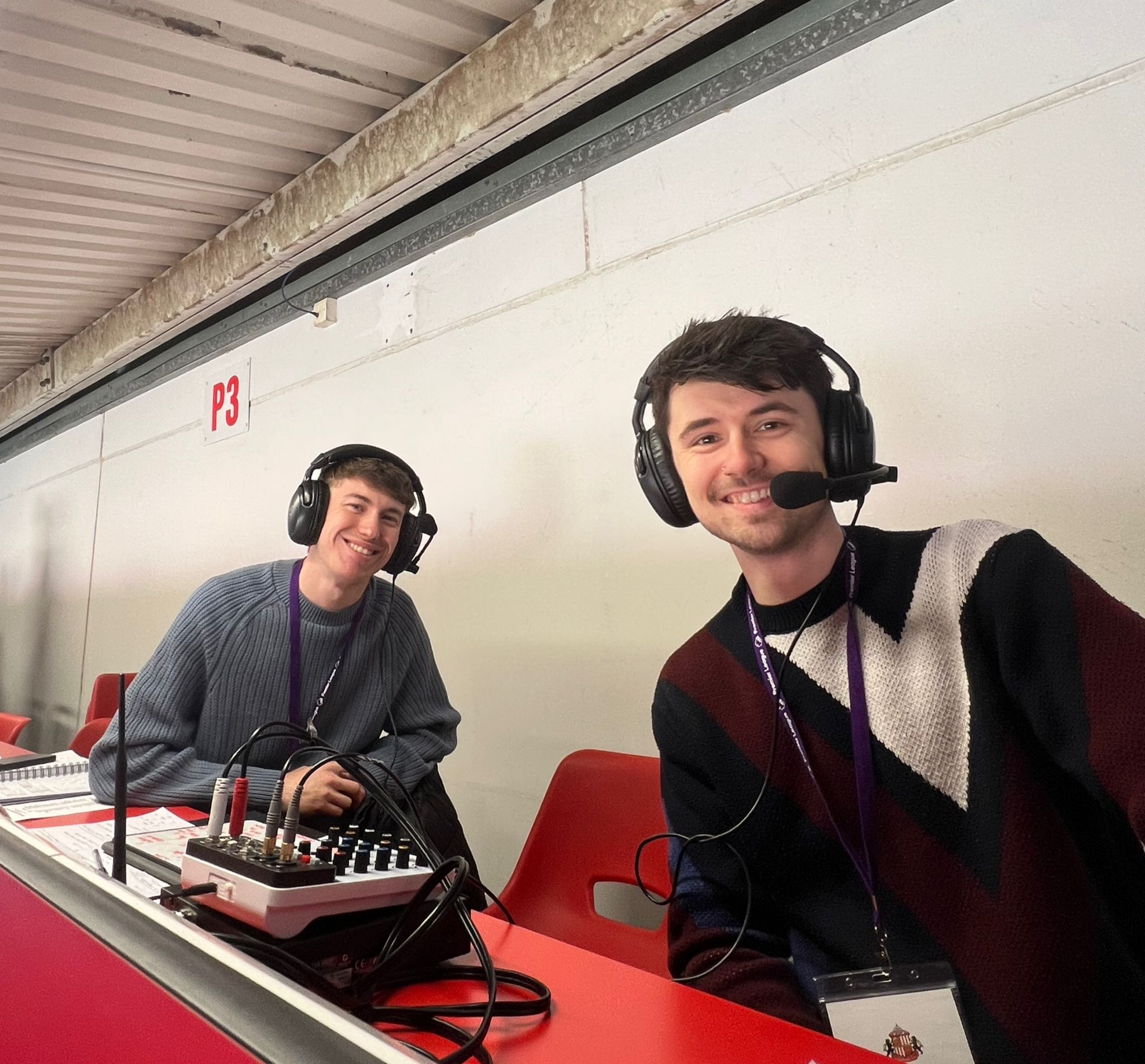 Twho commentators sit and pose for the camera from their comentary positio, with their backs to the back of the stand. 
