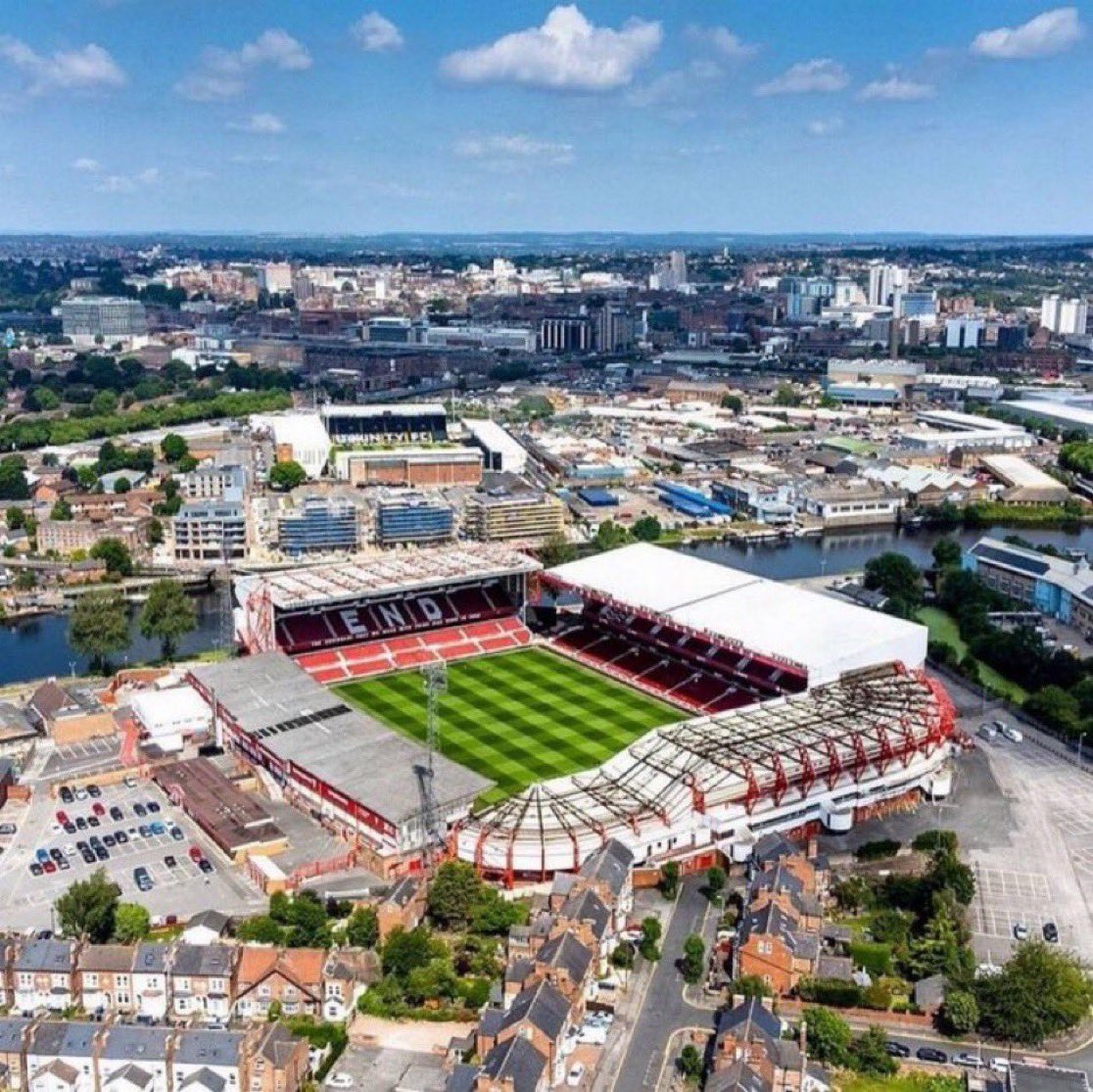 Aerial photo of Nottingham Forest’s City Ground taken on a bright spring day. The point of view is looking straight over the top of the adjoining Bridgford and Peter Taylor (Main) stands. The stadium is empty and the reds seats of the Trent End and Brian Clough stands are visible towards the back of the image. The pitch looks immaculate, a mixture of dark and light green squares making up the whole playing surface. At the very back of the photo the dark blue water of the River Trent can be seen running from left to right, horizontally.
The stadium is empty.