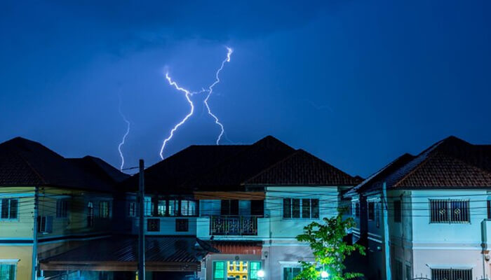 Lightning striking above residential homes at night, illustrating how lightning protection systems safeguard homes during severe storms.