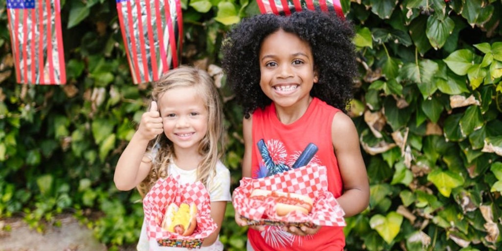 Smiling children enjoying hot dogs amidst an outdoor American flag celebration.