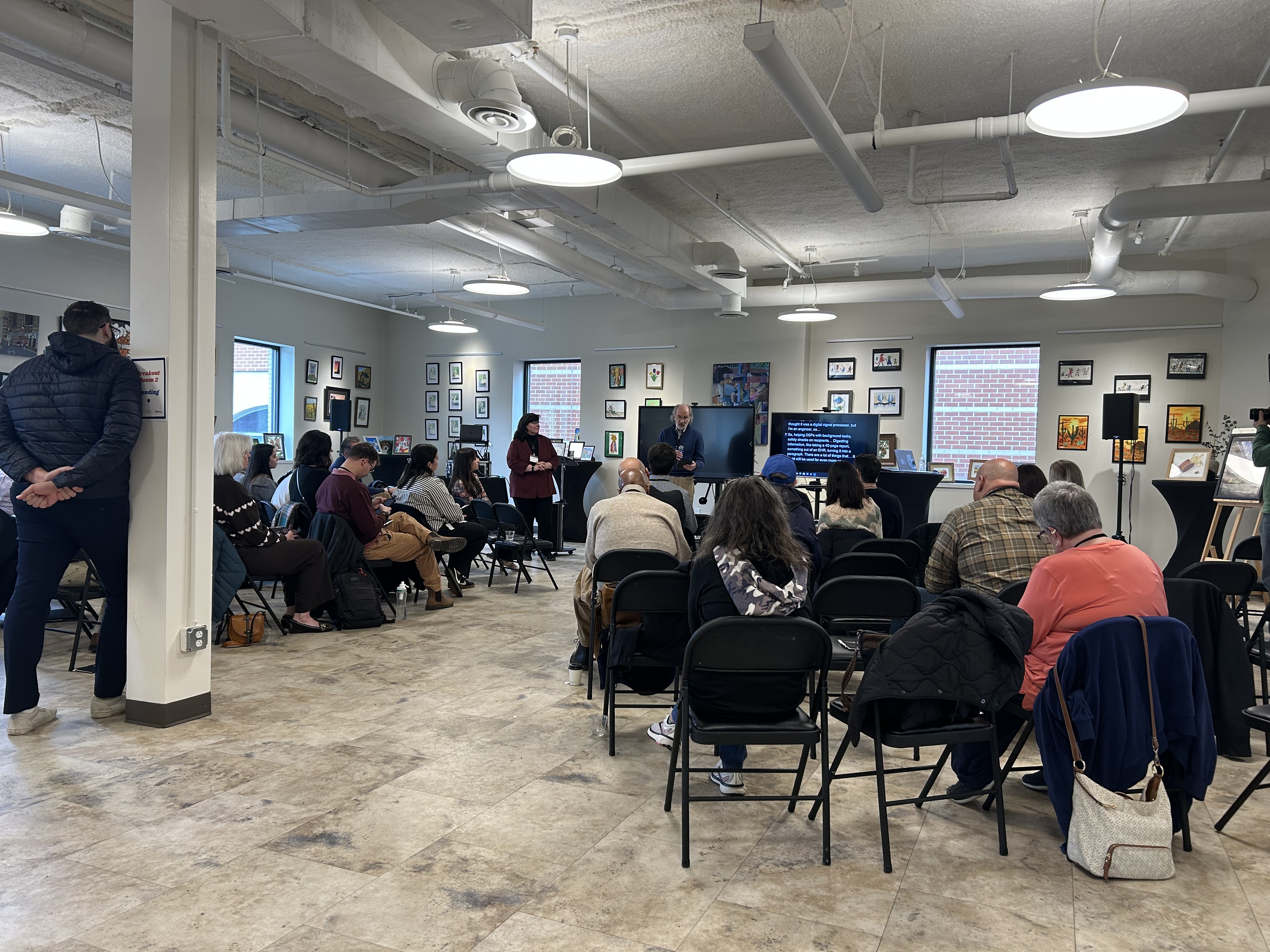 A group of attendees seated in chairs listening to a presenter speaking beside a screen.
