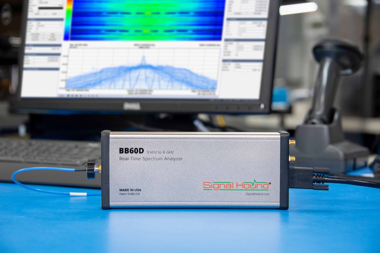 A Signal Hound BB60D Real-Time Spectrum Analyzer situated on a lab bench with a graphical analysis displayed on a screen in the background.