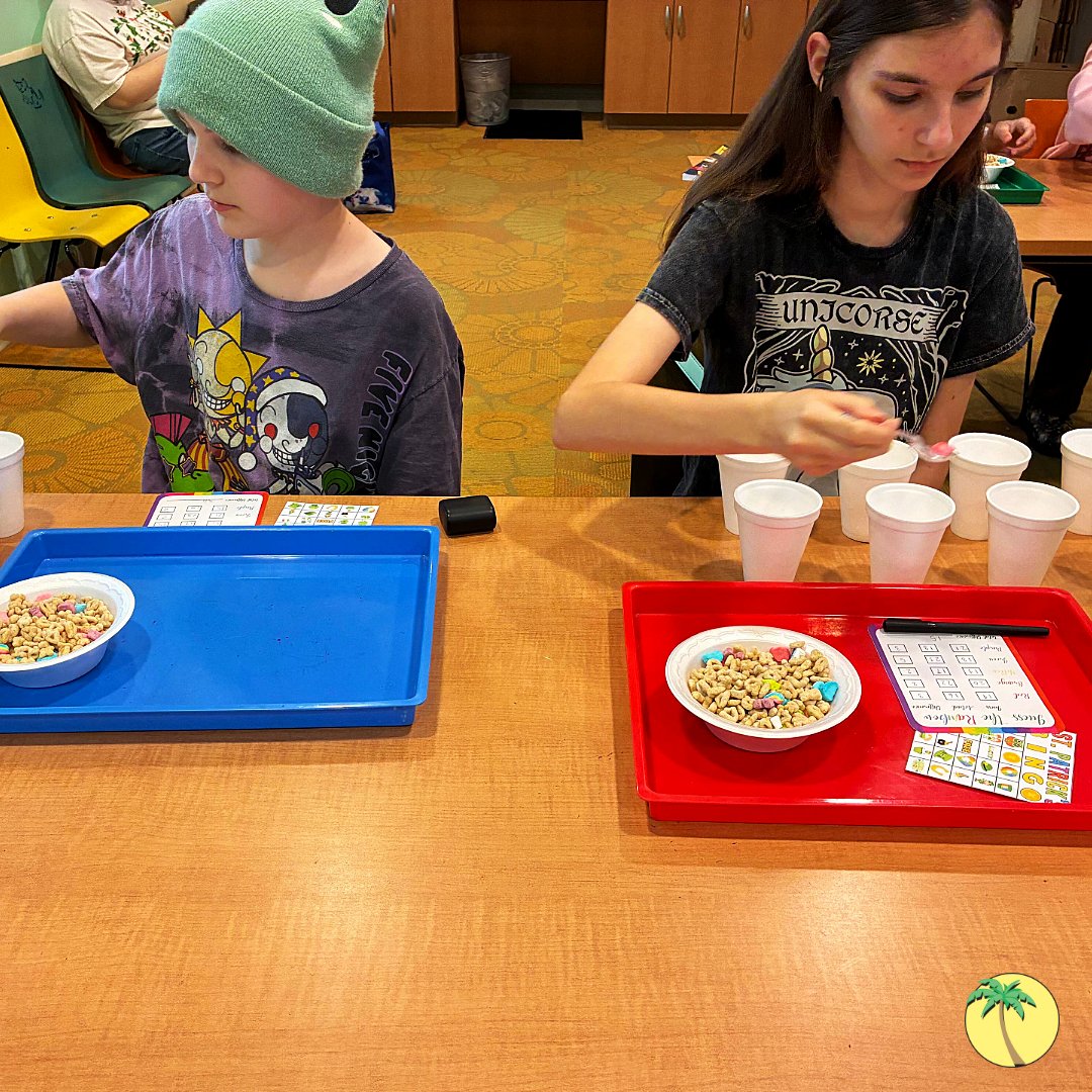 Two teenagers picking apart a bowl of Lucky Charms cereal and sorting the pieces into styrofoam cups. They have tally cards to measure how many of which charms they have.