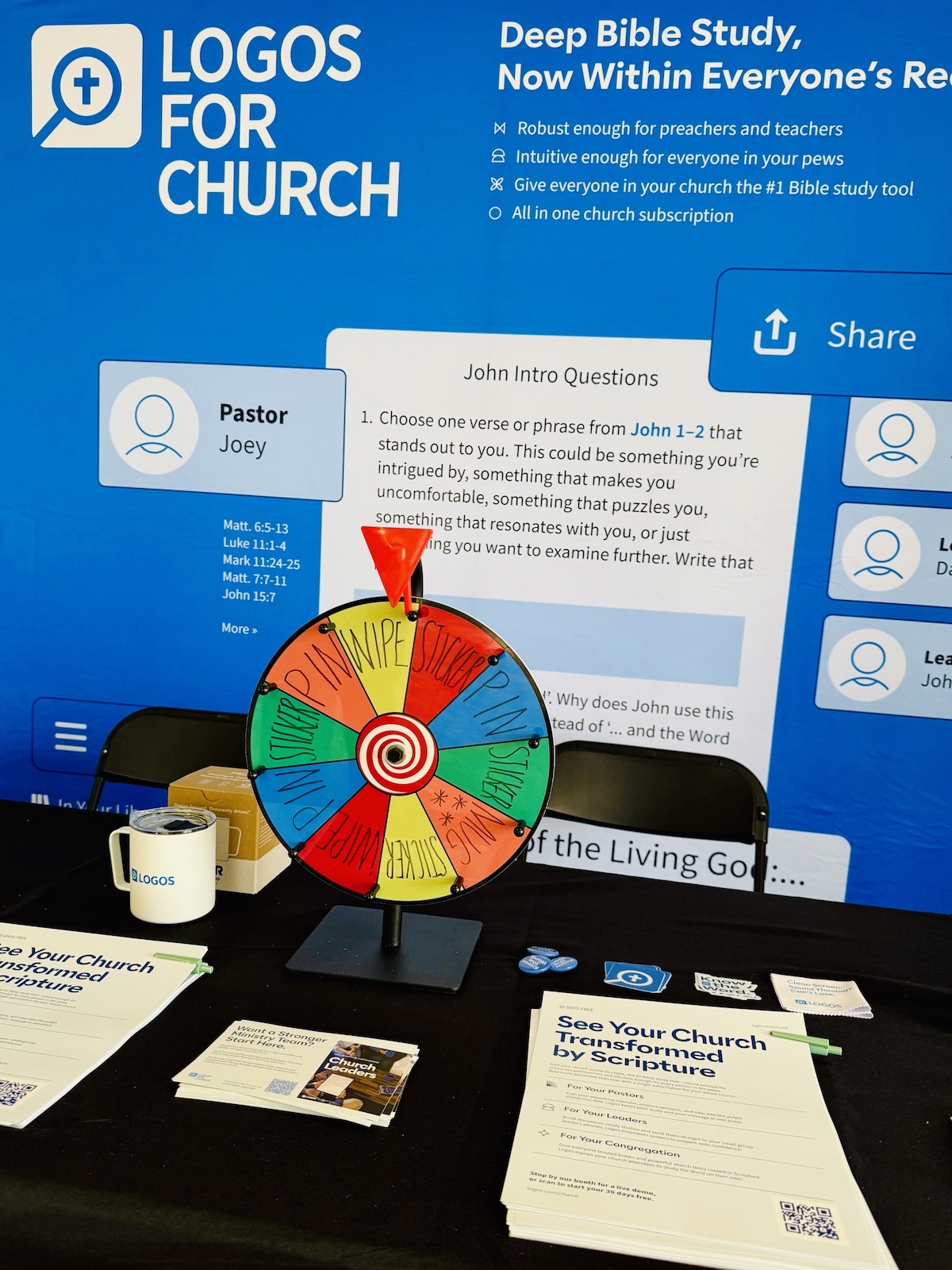 A table display for "Logos for Church" features a colorful spin wheel labeled with options like "Swipe," "Pass," and "Win." Behind it is a blue backdrop with text promoting deep Bible study, including a visible passage from John 1:1–2. There are brochures, a branded mug, and a sign identifying a person as "Pastor Joey.