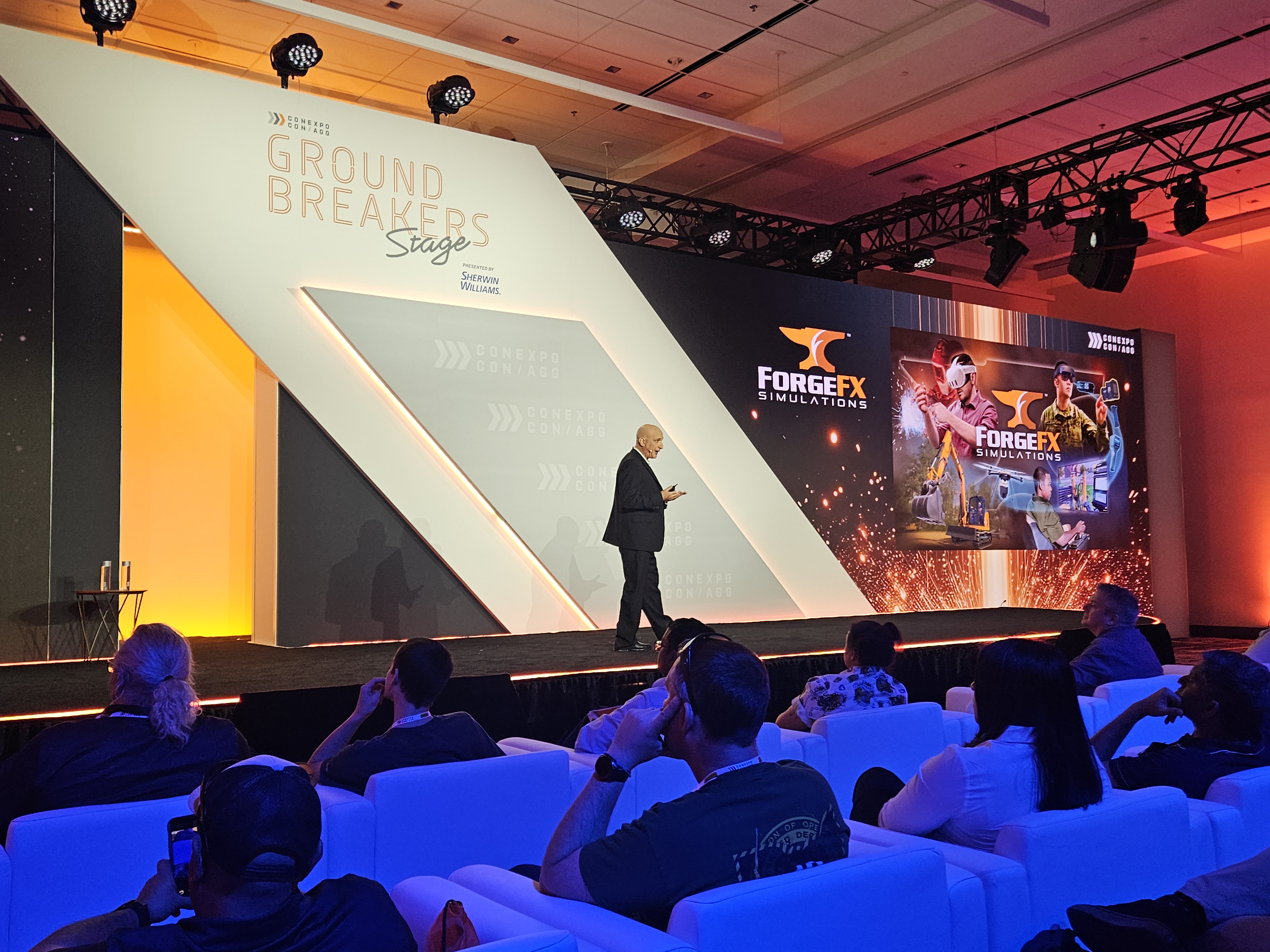 ForgeFX Simulations at CONEXPO-CON/AGG 2026: team members pose at the booth in front of a large video wall; CEO & Co-Founder Greg Meyers presents “From Iron to Impact” on the Ground Breakers Stage; attendees watch the session from white lounge seating; and visitors test immersive heavy equipment training demos using VR headsets and a bright orange lift simulator. Additional images show hands-on operator training, booth conversations, and live demonstrations of spatial computing-based training developed with partners including PICO XR. The photo set captures ForgeFX’s post-show highlights, customer engagement, and the company’s focus on safer, faster, and more measurable training for construction and industrial equipment operators.