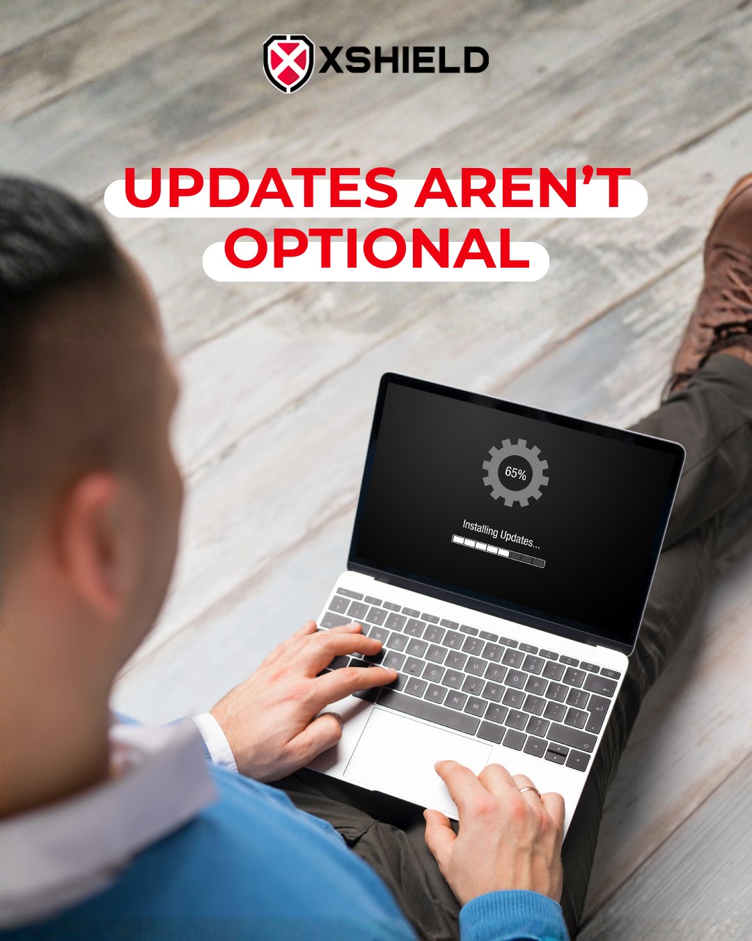 A person is sitting on the floor, using a laptop displaying a loading screen with "Installing Updates... 65%". Above, the text "UPDATES AREN'T OPTIONAL" is in bold red capital letters. The logo and name "XSHIELD" are at the top.