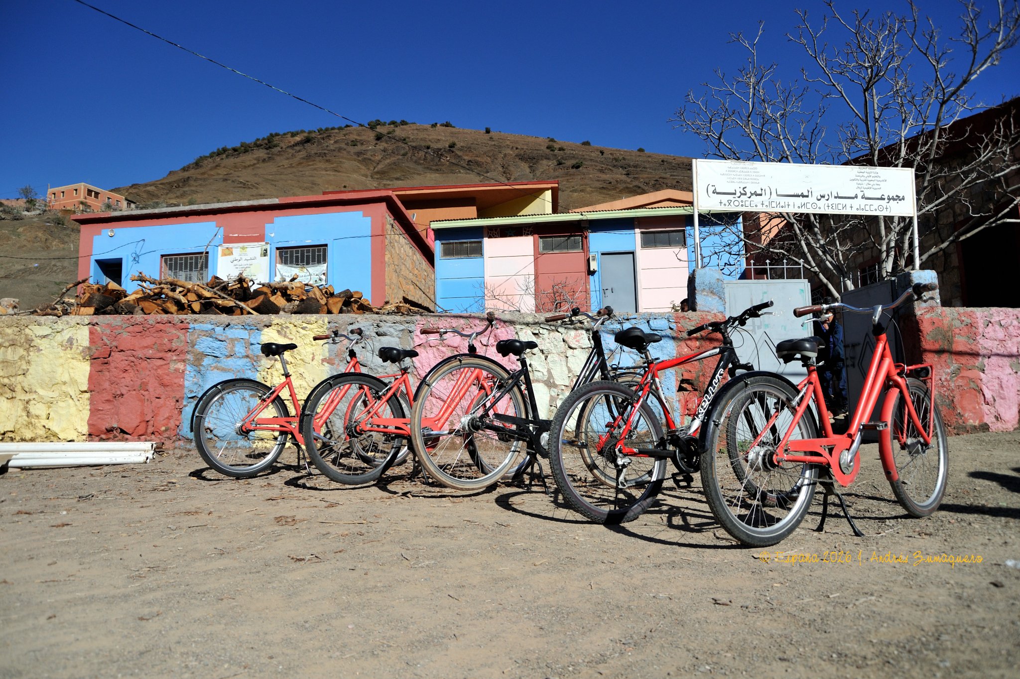 Cinco bicicletas esperan junto a un colegio. Las paredes del colegio están pintadas de diferentes colores. El cielo es de un azul intenso y el suelo que hay delante es de tierra.