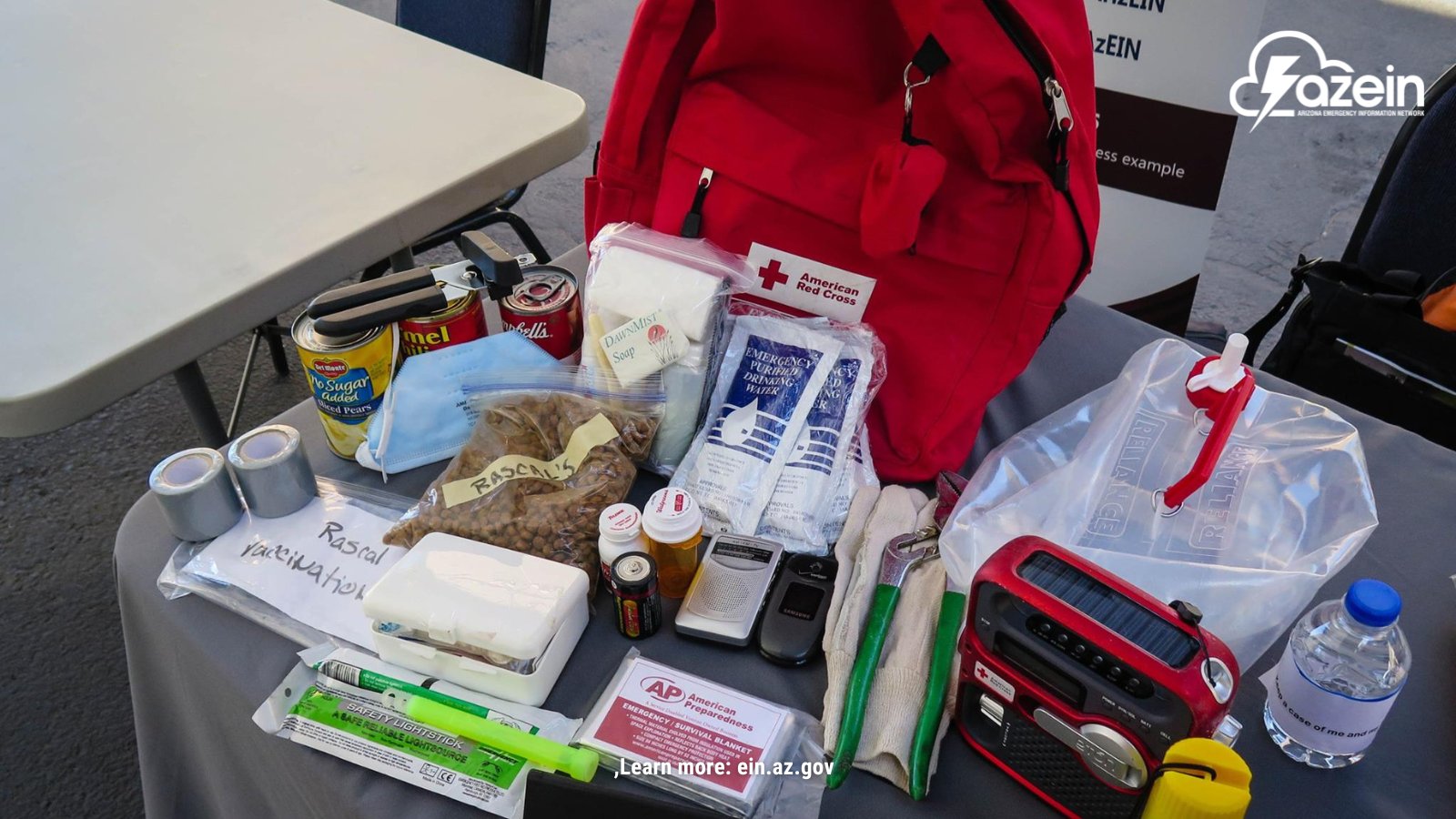 A flat lay photograph of various emergency supplies arranged on a grey tablecloth. Items include a large red backpack with an American Red Cross logo, canned food with a manual can opener, bags of pet food labeled "Rascal's," bottles of water, a solar-powered crank radio, duct tape, flashlights, a first aid kit, work gloves, and a document labeled "Rascal Vaccination." The "azein" logo is in the top right corner, and "Learn more: ein.az.gov" is centered at the bottom.