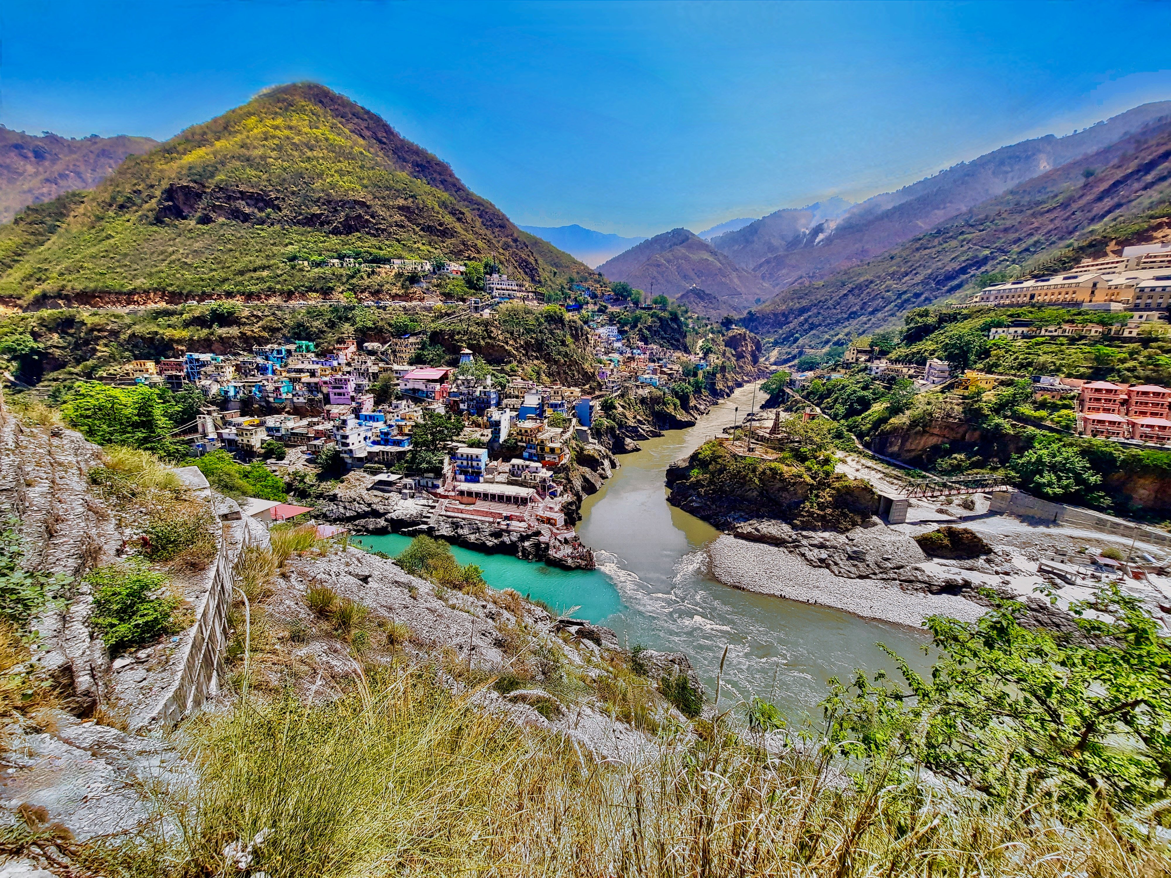 A breathtaking, wide–angle aerial view of the देवप्रयाग संगम in Uttarakhand. The vibrant turquoise अलकनंदा जी river meets the earthy–brown भागीरथी जी river to form the holy गंगा जी. The colorful houses of Devprayag town are perched on the steep mountain slopes under a clear blue sky. A sacred site of deep spiritual significance.