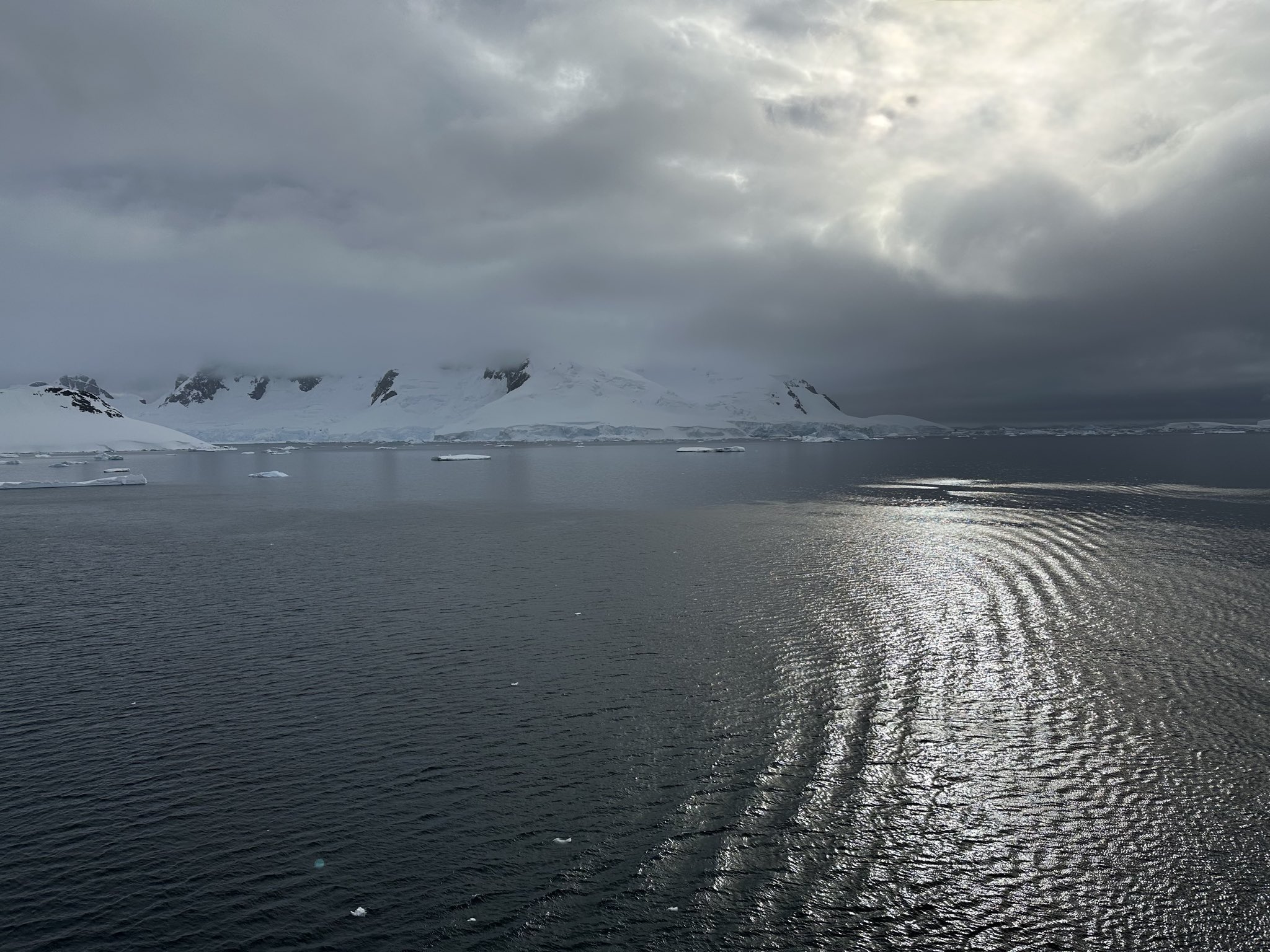 Dunkles Meer mit kleinen Eisschollen, dahinter schneebedeckte Berge unter dichten Wolken; ein Lichtstreifen spiegelt sich ruhig auf dem Wasser.
Die Szene wirkt still und fast zeitlos.